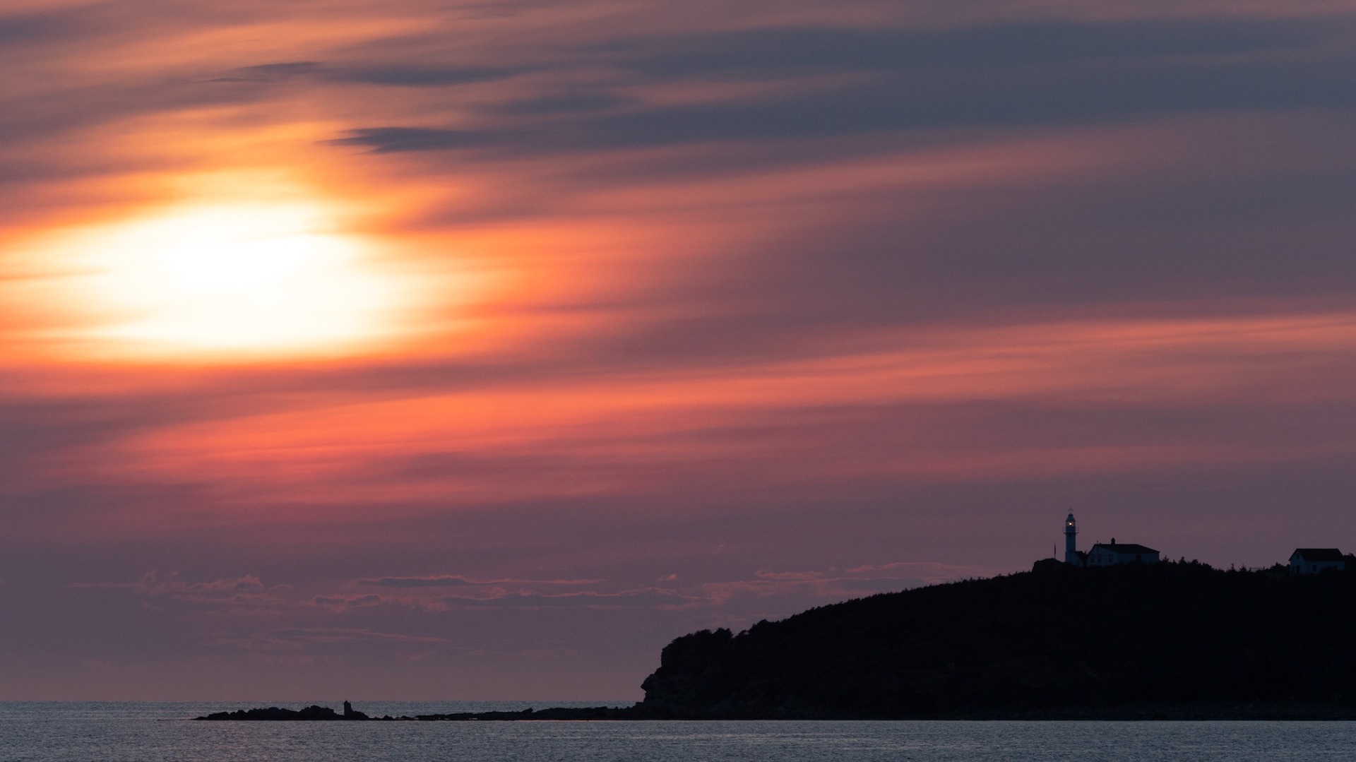 A mesmerizing Rocky Harbour sunset, where the sky is painted in soft streaks of orange, pink, and purple. The silhouette of Lobster Cove Lighthouse stands in the distance, quietly watching over the rugged coastline. As the sun sinks below the horizon, its golden light reflects across the water, creating a scene of tranquility and timeless coastal beauty.