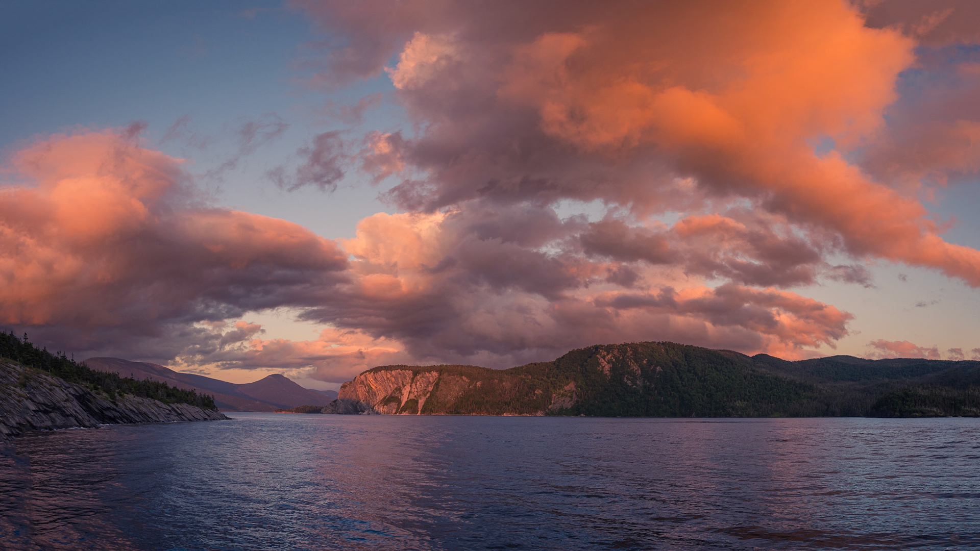 A breathtaking sunset over Norris Point, as seen from the Cat Stop Pub, where the evening sky ignites in hues of pink and gold. The dramatic clouds reflect the fading light, casting a warm glow over Bonne Bay. In the distance, the rugged face of Shag Cliff stands tall, a timeless sentinel against the vast Newfoundland wilderness. A perfect scene of coastal serenity and natural wonder.