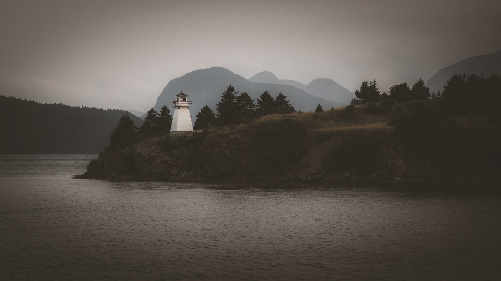 A moody, atmospheric view of the Woody Point Lighthouse, standing as a solitary beacon against the misty mountains of Gros Morne National Park. Perched on a rugged coastline, this iconic lighthouse has guided mariners through the waters of Bonne Bay for generations. The soft, diffused light and dramatic landscape capture the quiet resilience and beauty of Newfoundland’s coastal heritage