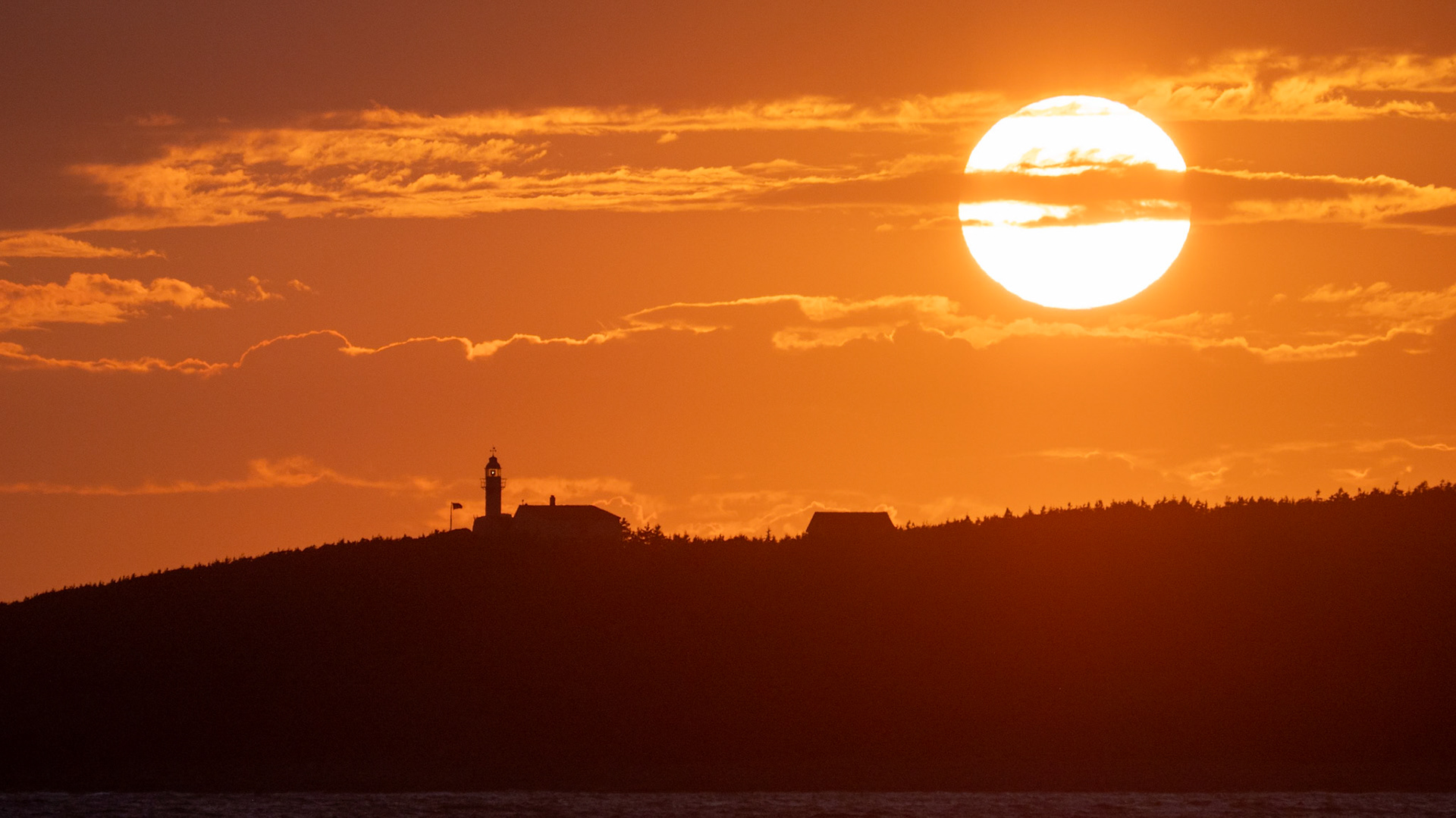 A fiery sunset over Rocky Harbour, casting an ethereal glow across the sky as the silhouette of Lobster Cove Lighthouse stands in quiet defiance against the fading light. The vast sun, veiled by delicate streaks of cloud, creates a mesmerizing contrast between warmth and shadow, capturing the essence of Newfoundland’s rugged beauty and maritime heritage.