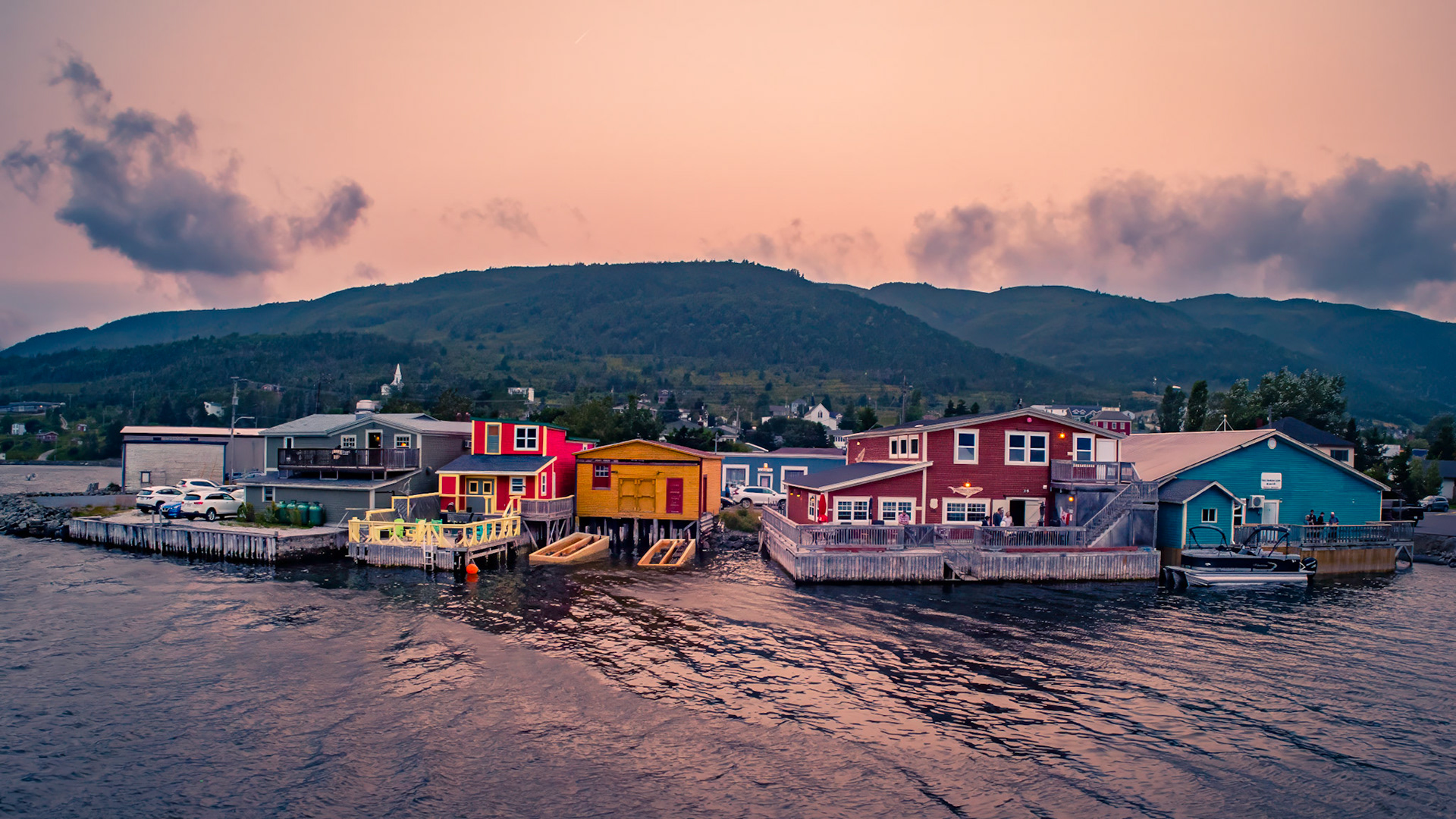 A vibrant evening in Woody Point, where colorful waterfront buildings stand against the lush backdrop of the Long Range Mountains. As the warm hues of sunset cast a soft glow over the town, the quiet ripples on the water reflect the charm and character of this coastal gem in Gros Morne National Park. A perfect blend of heritage, community, and stunning Newfoundland scenery.