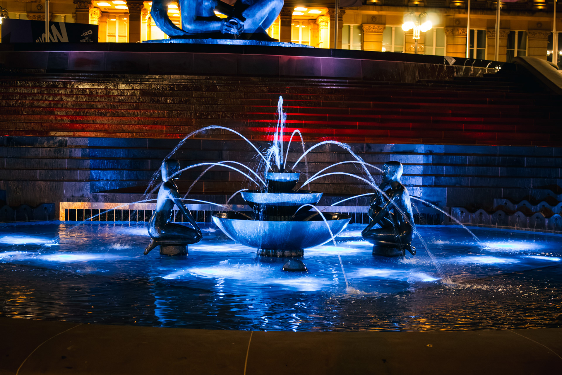 A photo of a fountain in birmingham lit up at night with red and blue lights.
