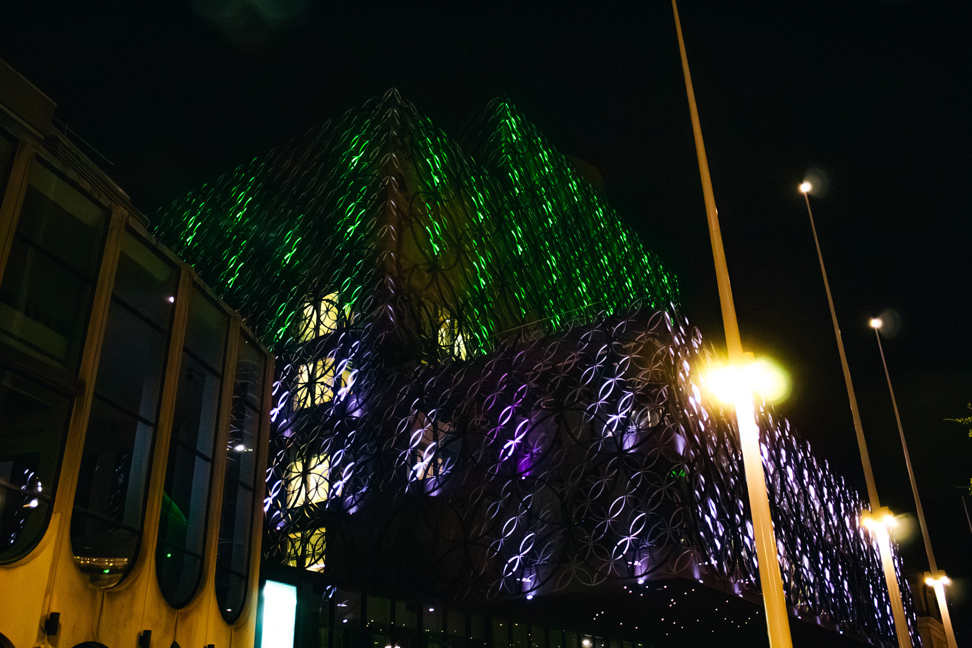 A photo of birmingham library lit up at night in green and purple lights.