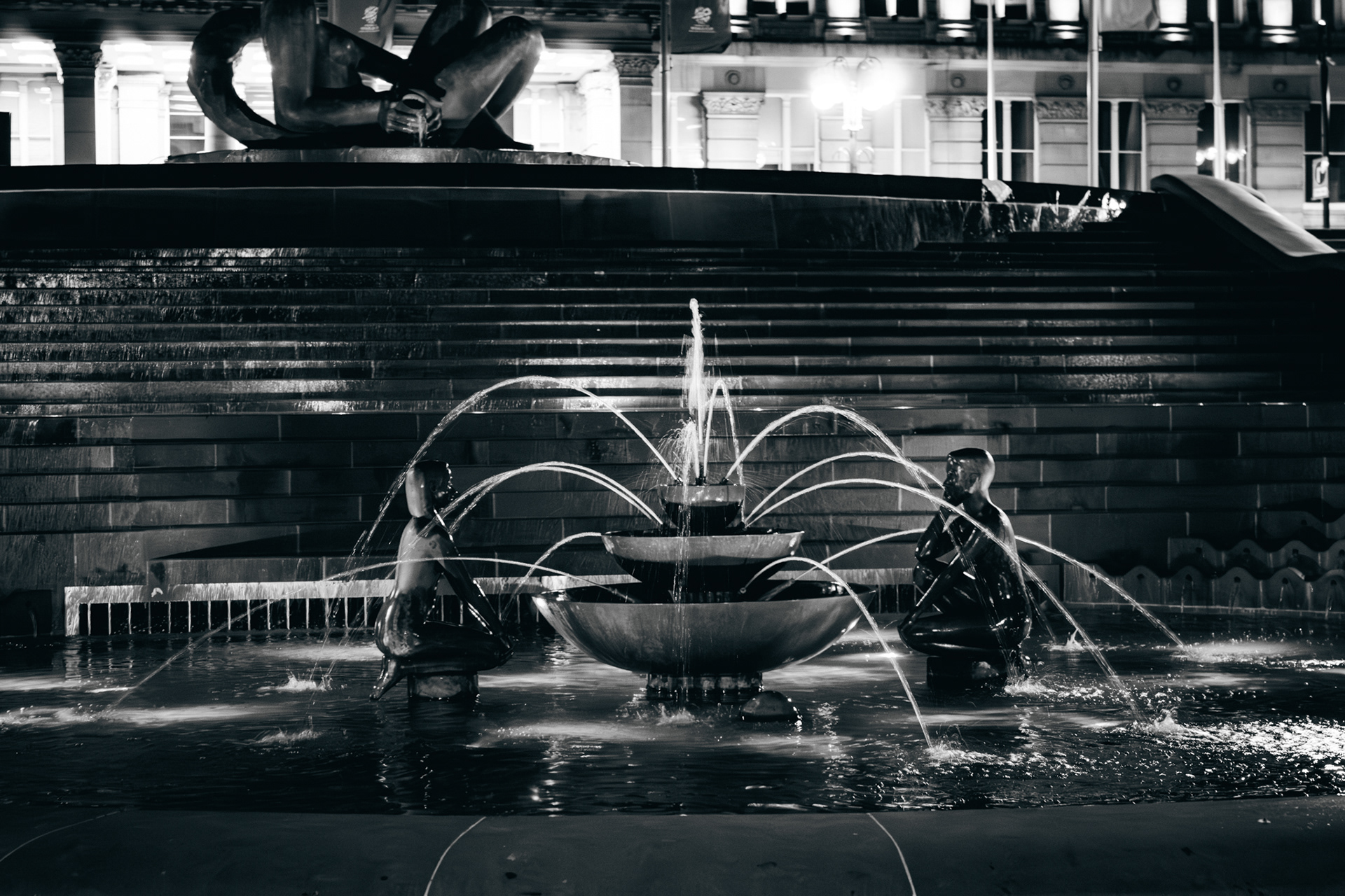 A black and white photo of a fountain in birmingham lit up at night.