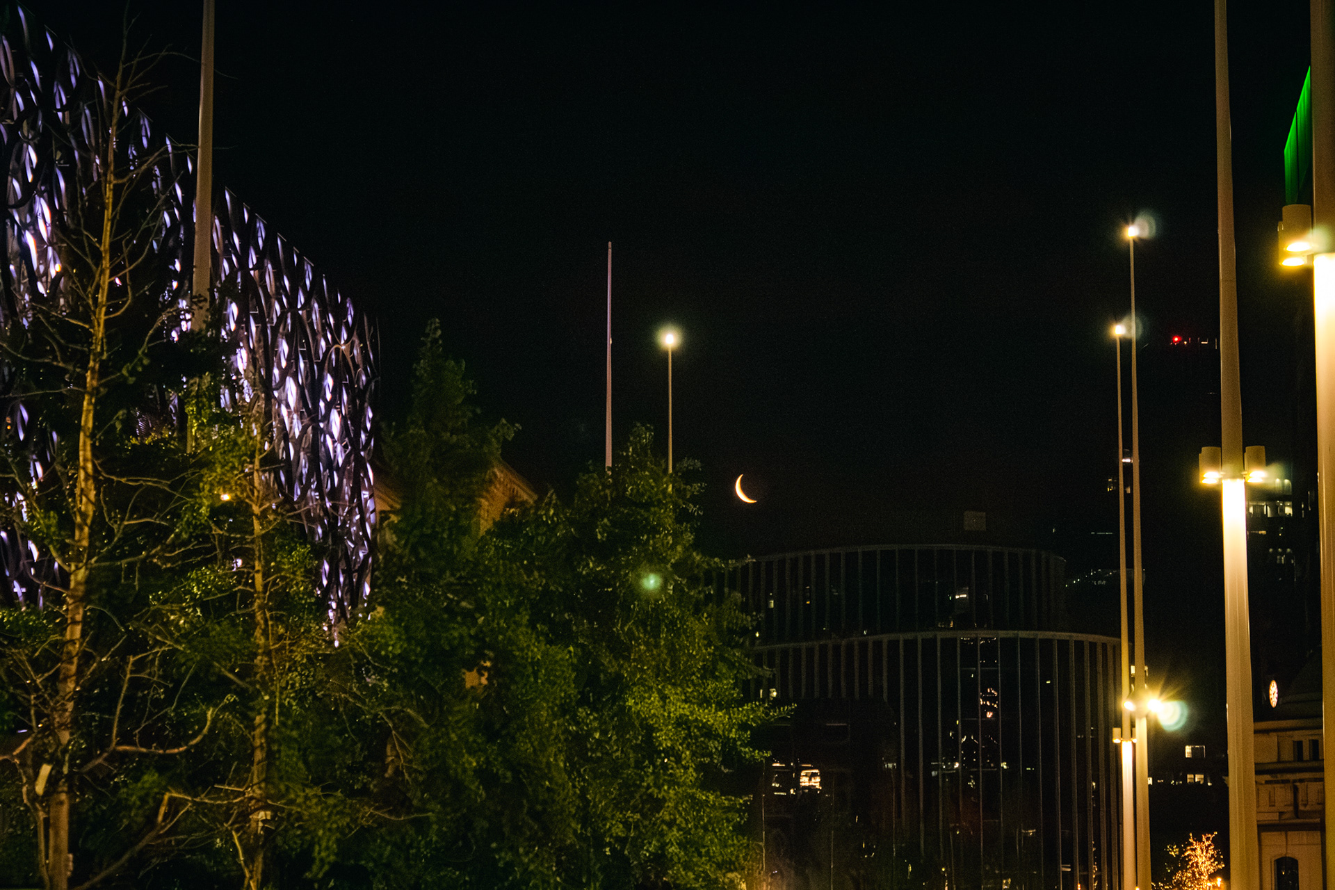 A picture of birmingham library lit up at night with the moon visible in a crescent shape.