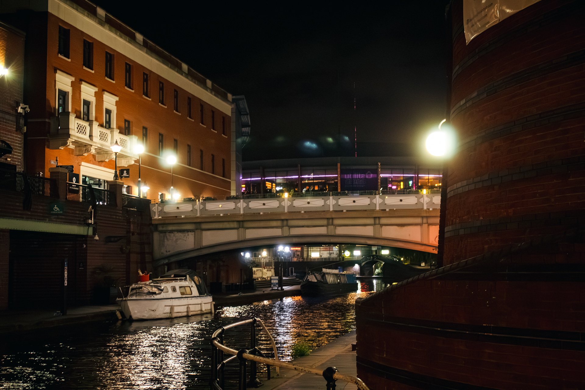 A night time shot of a bridge across a canal with a small boat on the left of the screen.