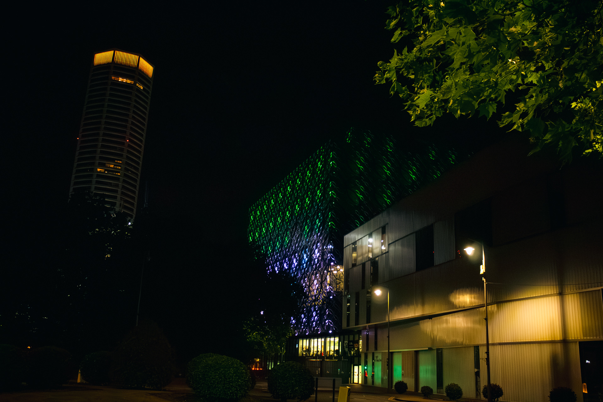 A photo of birmingham library lit up at night in purple and green with the octagon building on the left of the screen and the back of the rep theatre next to the library