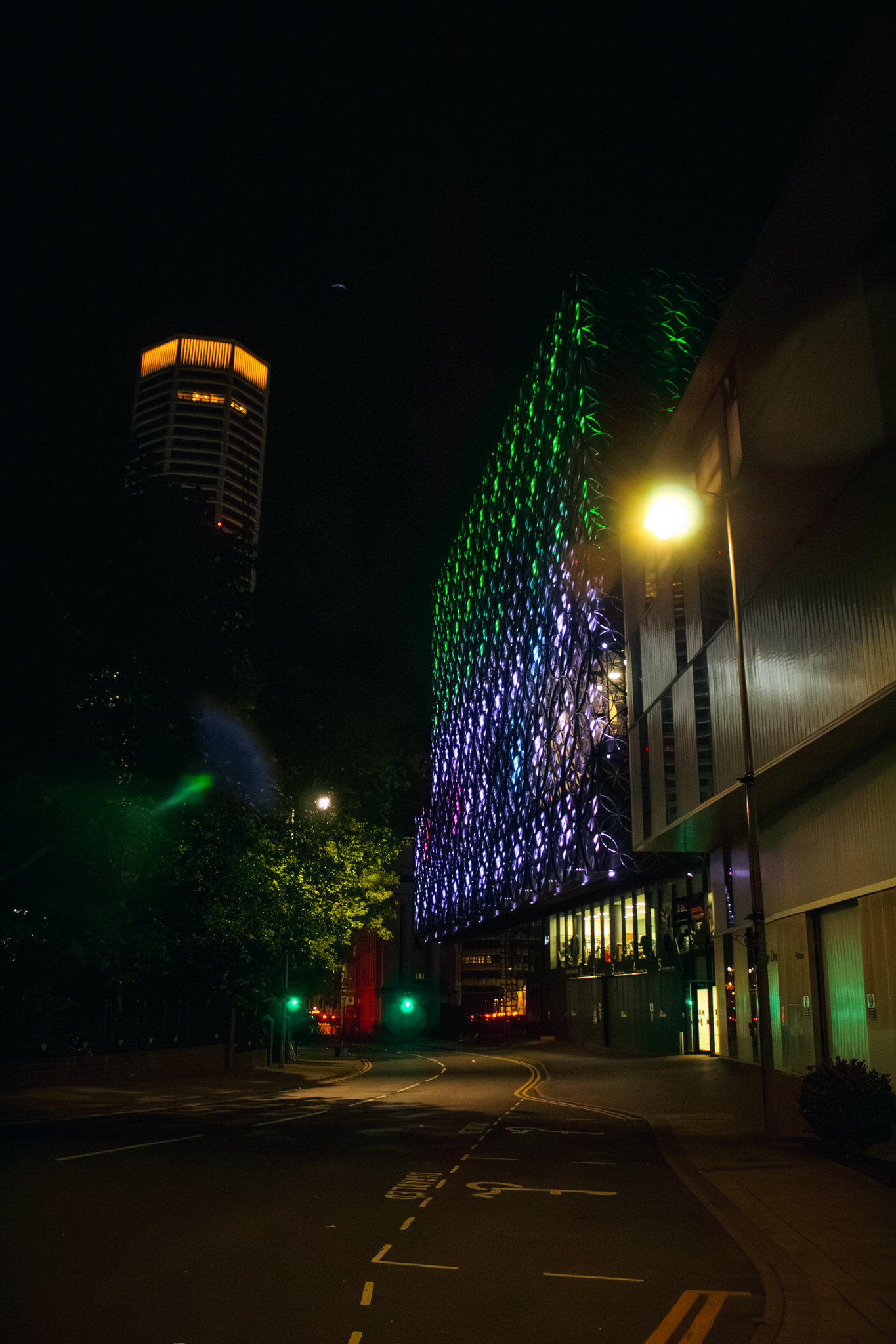 A vertical photo of birmingham library lit up at night in purple and green with the octagon building on the left side of the screen.