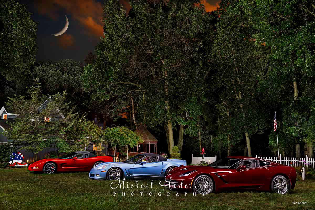 A pretty light-painted photograph of  three Corvettes. A Torch Red C5 2002 Corvette Convertible, a 2012 Carlisle Blue Corvette Grand Sport Convertible, and a Long Beach Red 2019 Corvette Stingray Coupe.