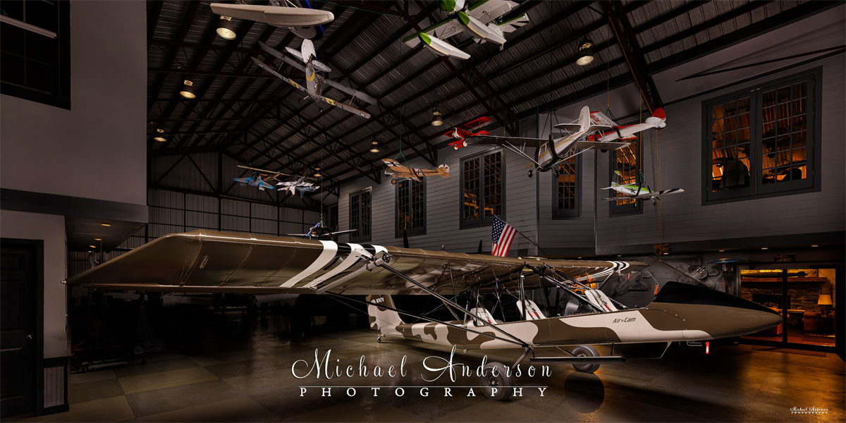 A cool light painted photograph of AirCam N74TV in a hangar filled with remote-controlled aircraft suspended from the ceiling.