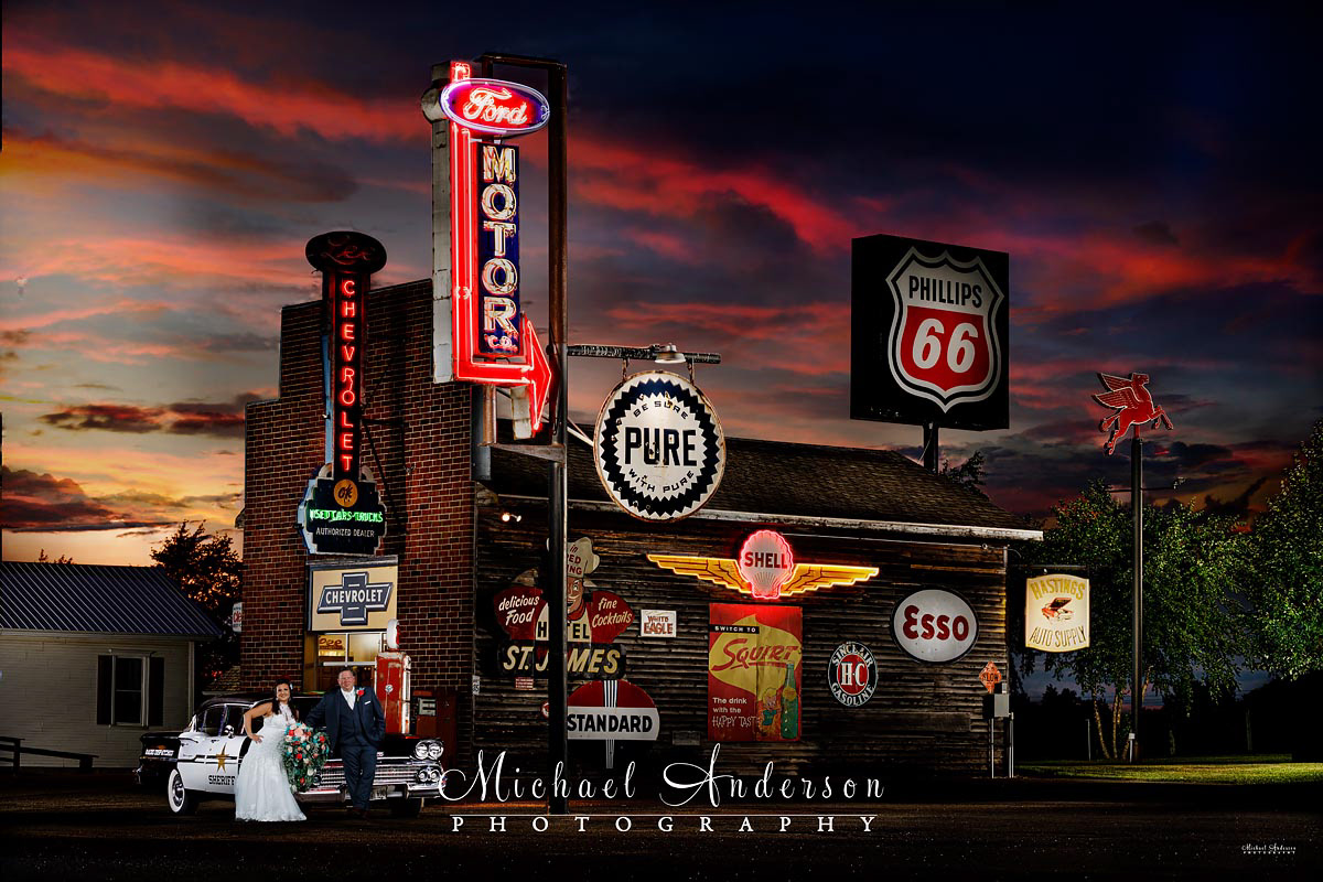 A cool light painted wedding photograph of a bride and groom, by a 1950's Sheriff's Car and Chevy Dealership, at Little Log House Pioneer Village in Hasting, MN.