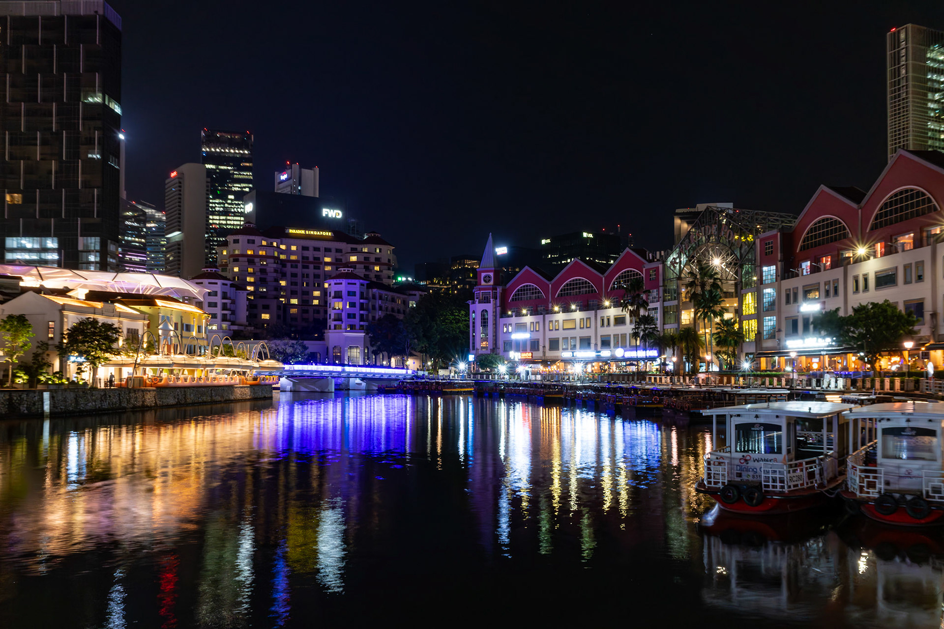 Spaziergang entlang des Flusses Richtung Hafen; Lichtspuren und beleuchtete Gebäude begleiten den Weg.
