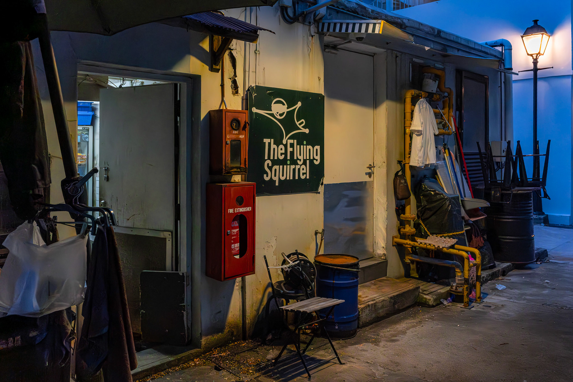 Kleine Gasse mit lokalem Flair bei Dämmerung; enge Bebauung mit typischen Ladenfronten und Beschilderung.