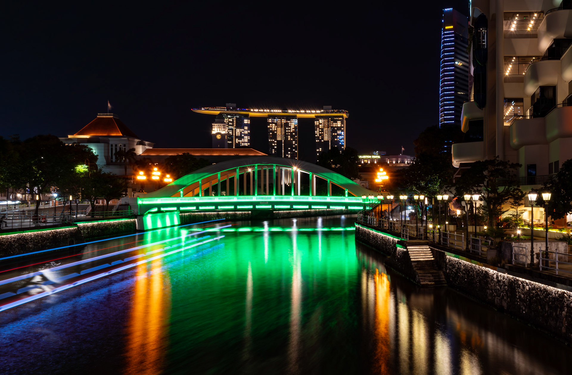 Flusspromenade mit Beleuchtung und Brücke; reflektierende Lichter im Wasser und belebte Uferbereiche. Lichtspur eines Schiffs.