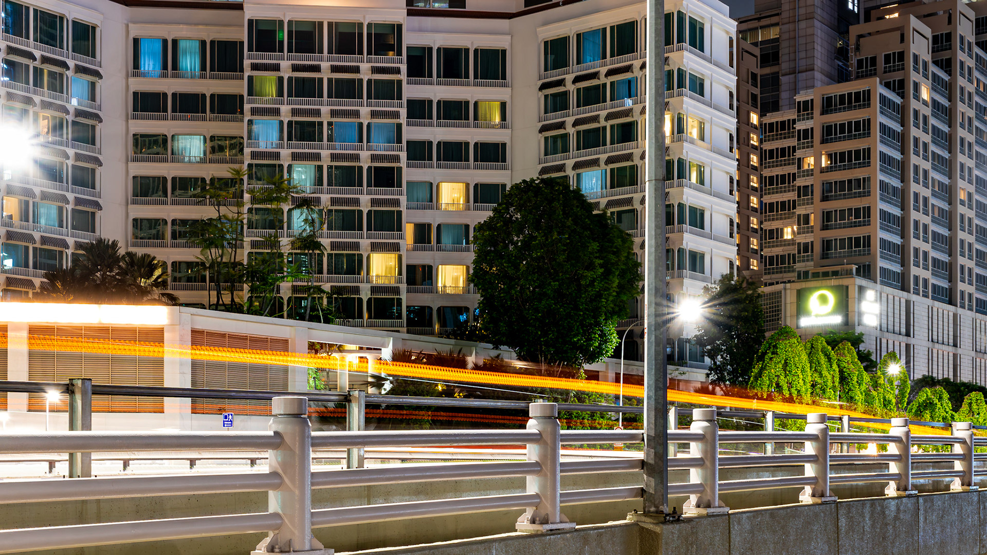 Orangefarbene Lichtspuren hinter einem weißen Ufergeländer. Der Hintergrund besteht vollständig aus der gerasterten Fassade eines Hochhauskomplexes.