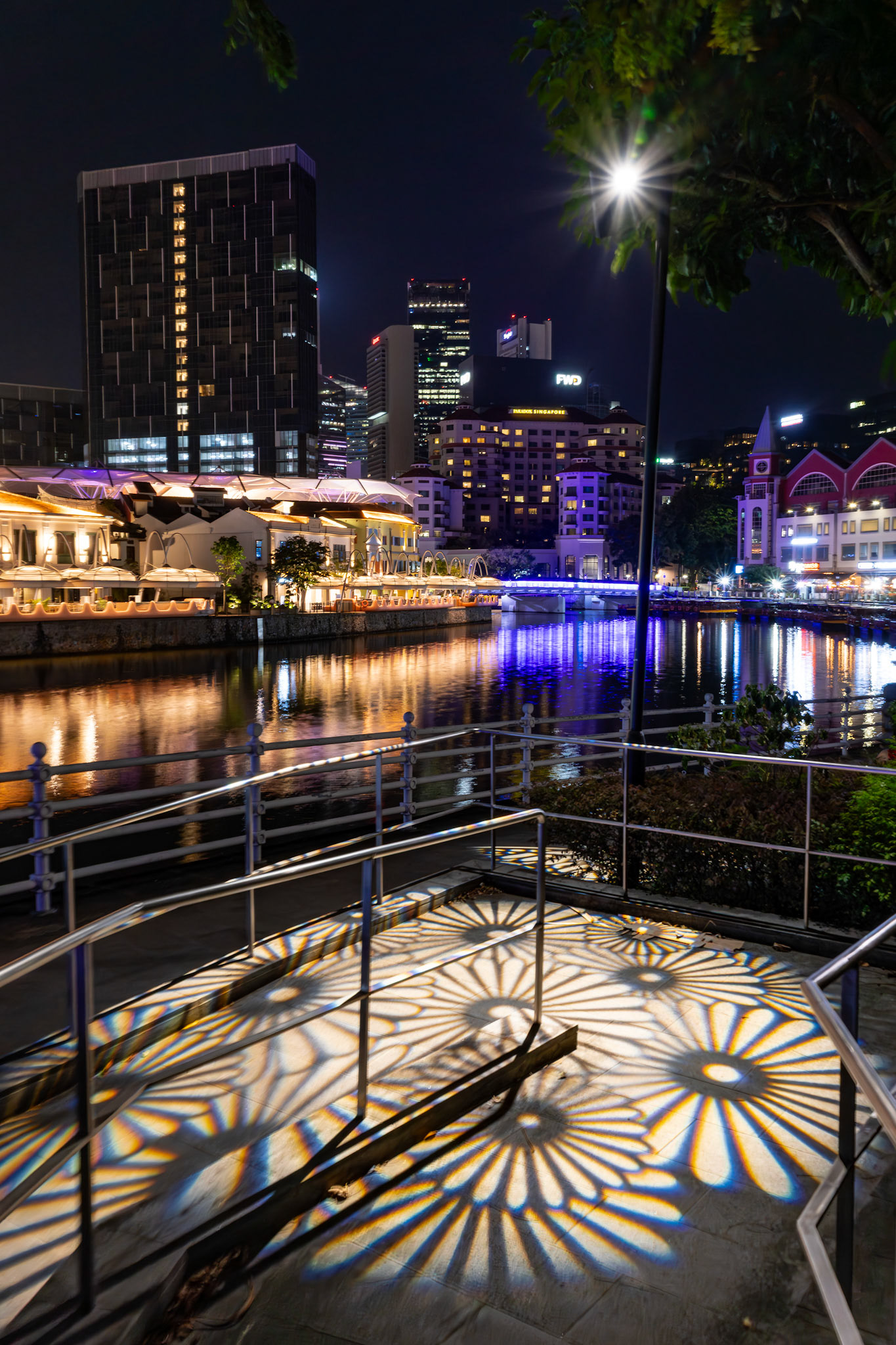 Eine Laterne wirft ein geometrisches Lichtmuster auf das Uferpflaster. Im Hintergrund die touristisch illuminierte Skyline.