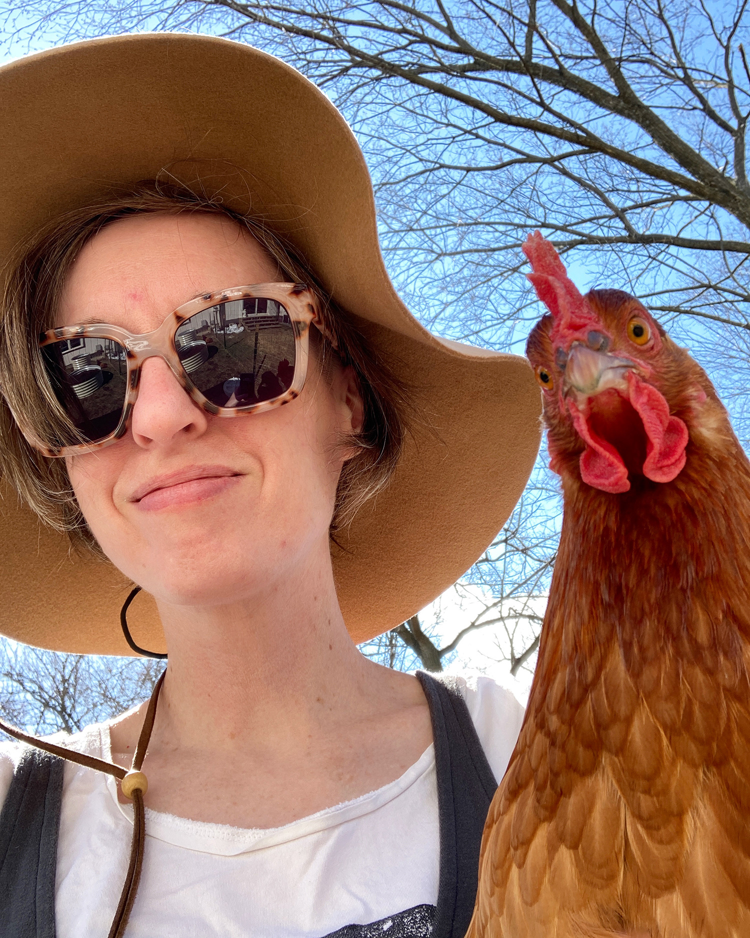 smiling woman wearing a sunhat and glasses, holding a chicken on a sunny day