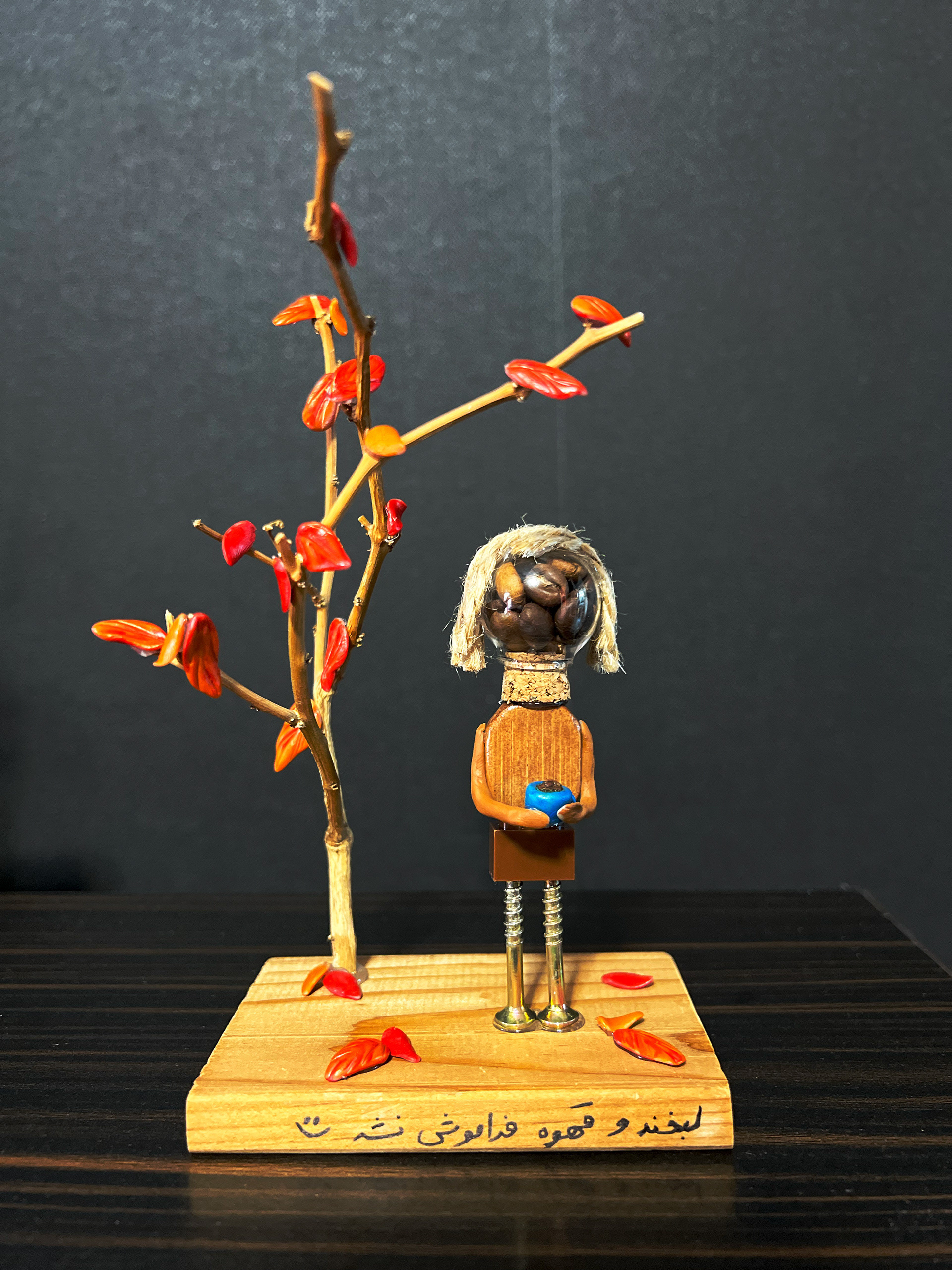 A tactile portrait of a coffee enthusiast and nature lover. Crafted to mirror her essence, the figure’s wooden body grounds her in the earth, while her head, crowned with rich coffee beans, celebrates the quiet joy of her daily ritual. Holding her favorite blue mug against a warm, autumn-themed backdrop, this handcrafted piece captures the cozy spirit of a crisp fall morning.