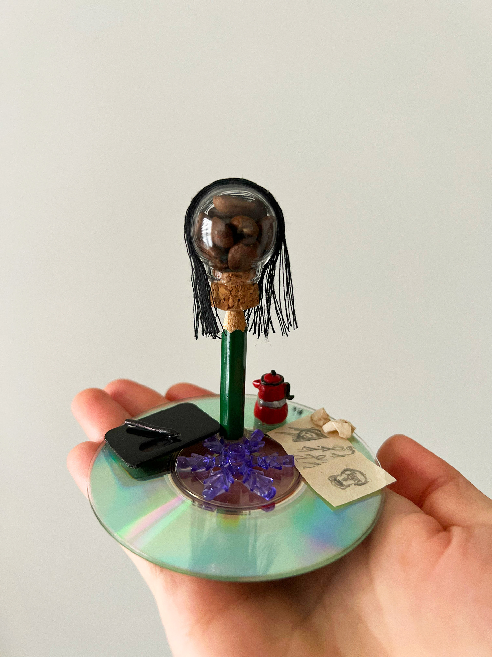 A sculptural celebration of resilience, art, and the tech that bridges them. Built with a drawing-pencil body and paired with a digital tablet, this figure honors her tireless dedication to her craft. A compact CD base roots her story in a computer background, while a head filled with coffee beans and a signature red coffee maker symbolize the warmth and creative energy she pours into her work. At its core, a delicate snowflake element honors her February birth, a quiet nod to a calm, brilliant spirit who meets every challenge with grace.