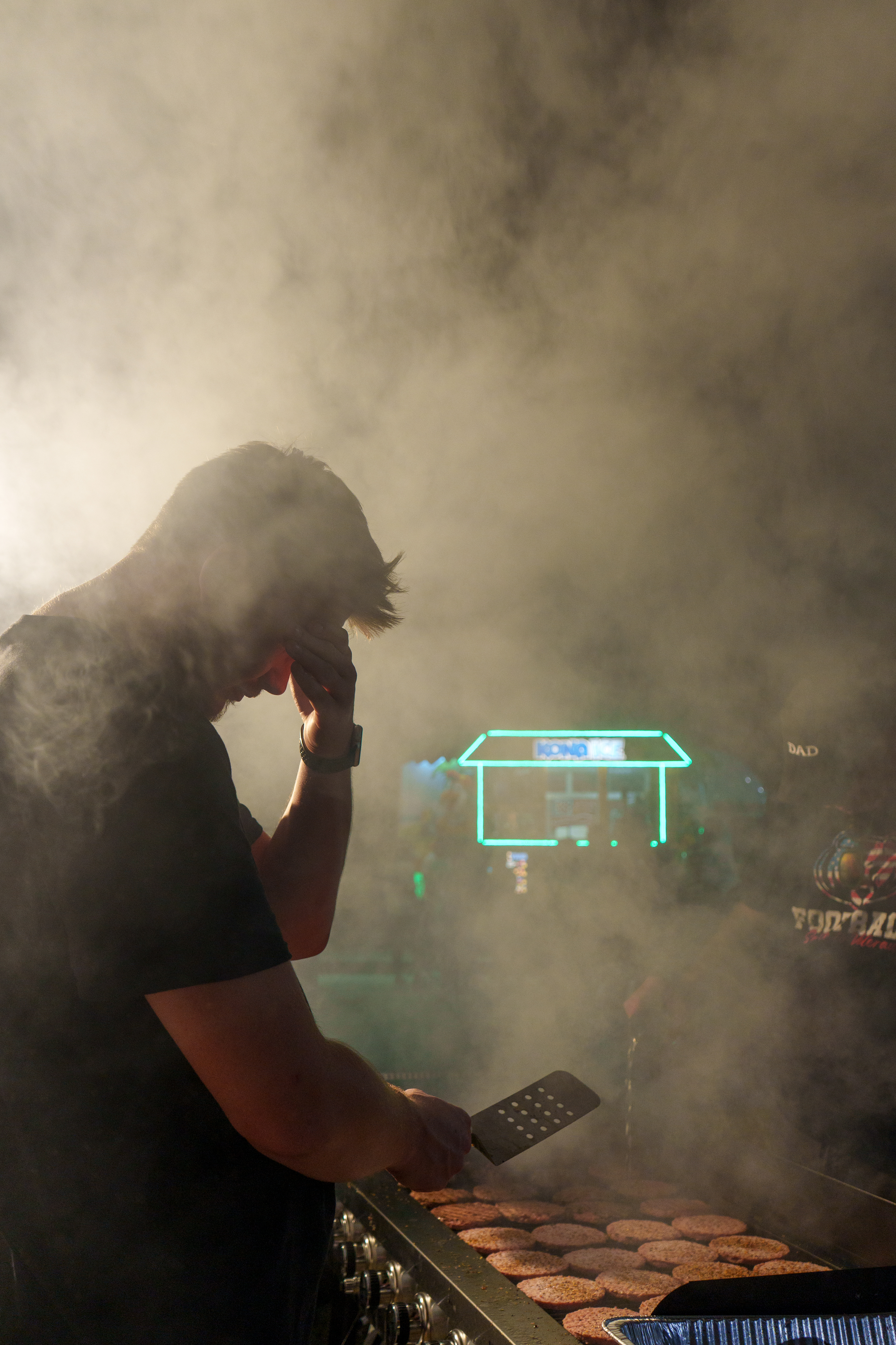 Ross Hagan shields his eyes from smoke rising from the grill as he cooks hamburgers during the Rock Bridge-Park Hill game on Aug. 29, 2025 at Rock Bridge High School in Columbia, Mo. Hagan is the boyfriend of Nicole Ream, whose family has run the Rock Bridge hamburger grill since 2021.