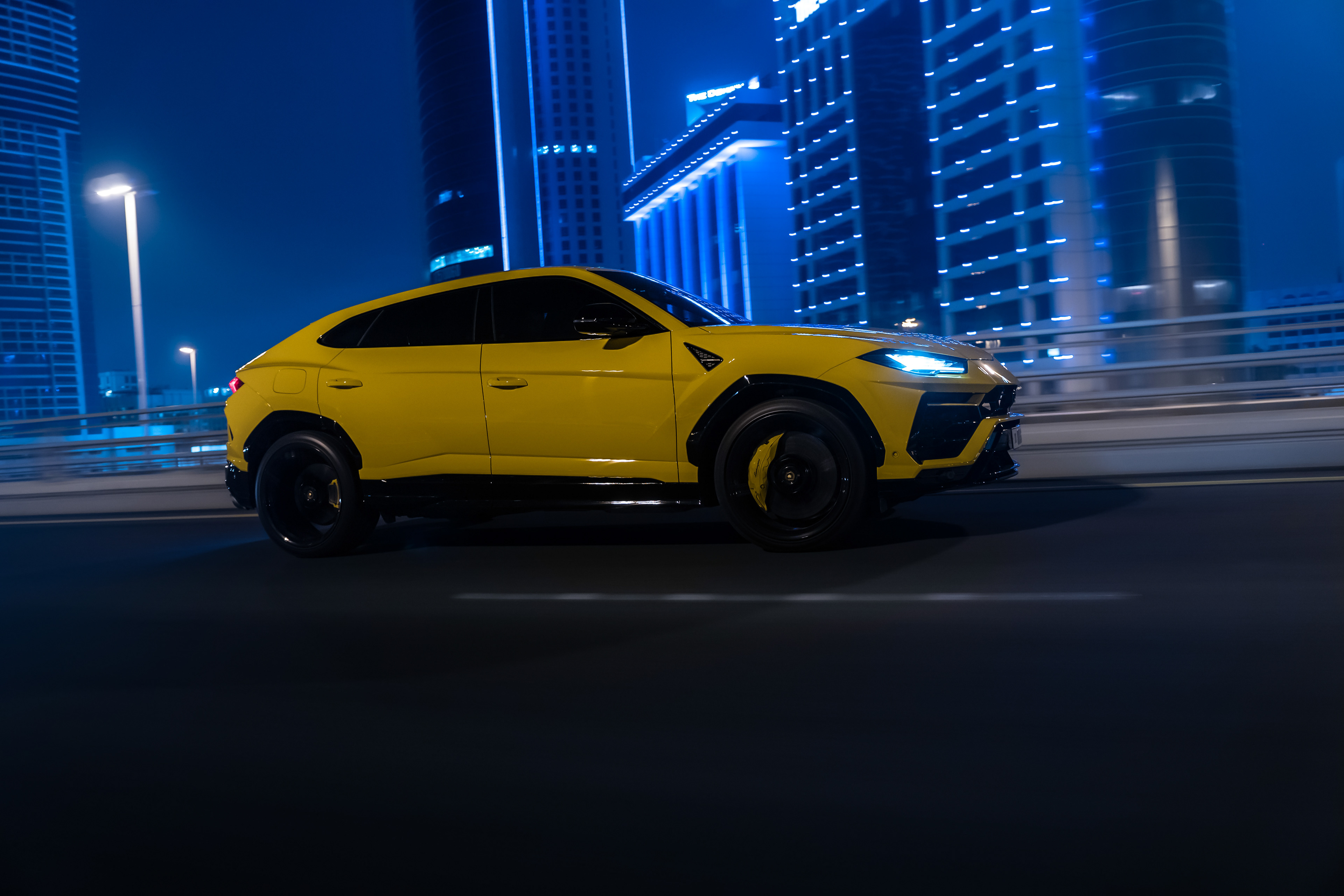 Side profile of a yellow Lamborghini Urus cruising at night through Bahrain’s illuminated skyline