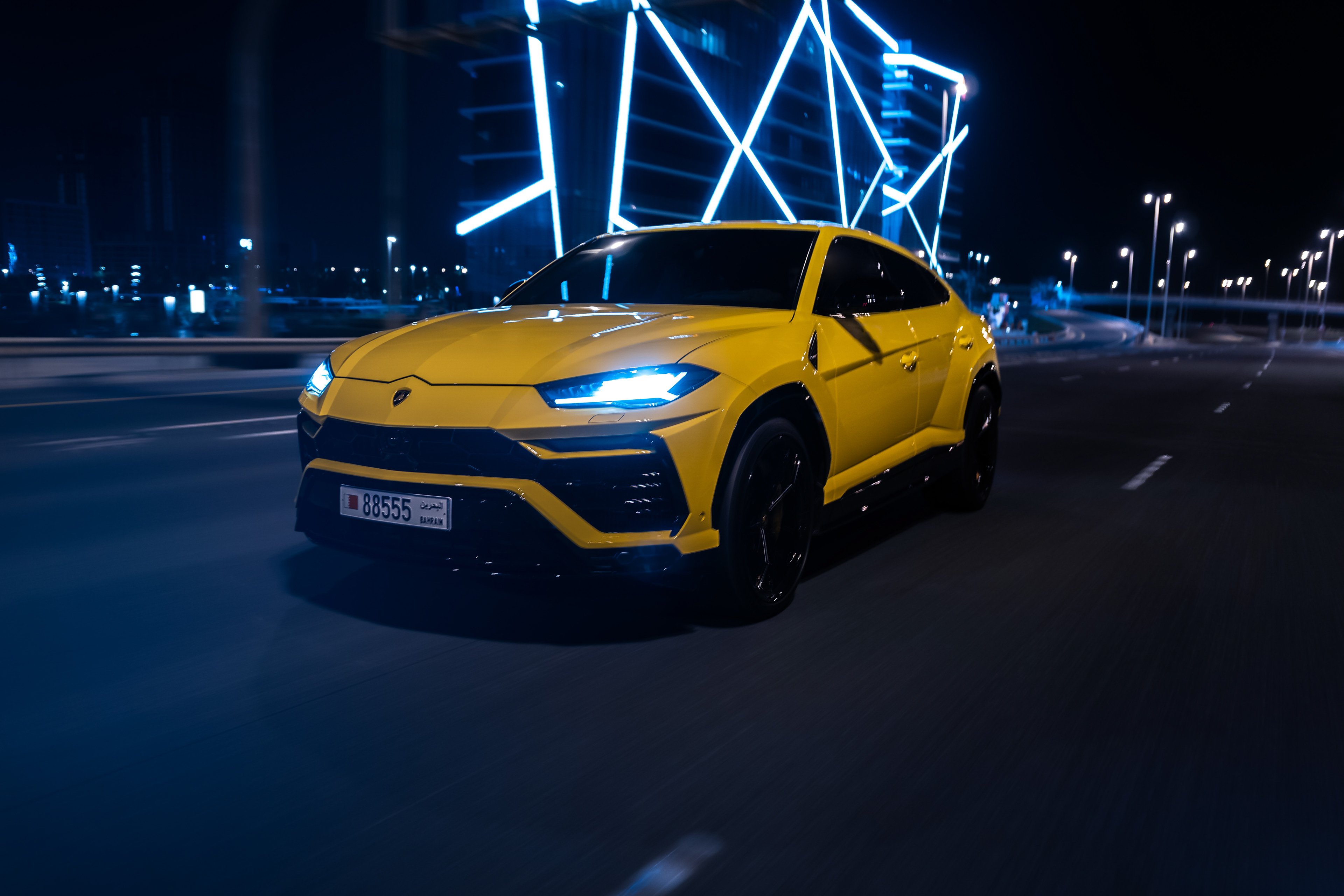 Front view of a yellow Lamborghini Urus in motion under Bahrain Bay’s glowing blue architecture