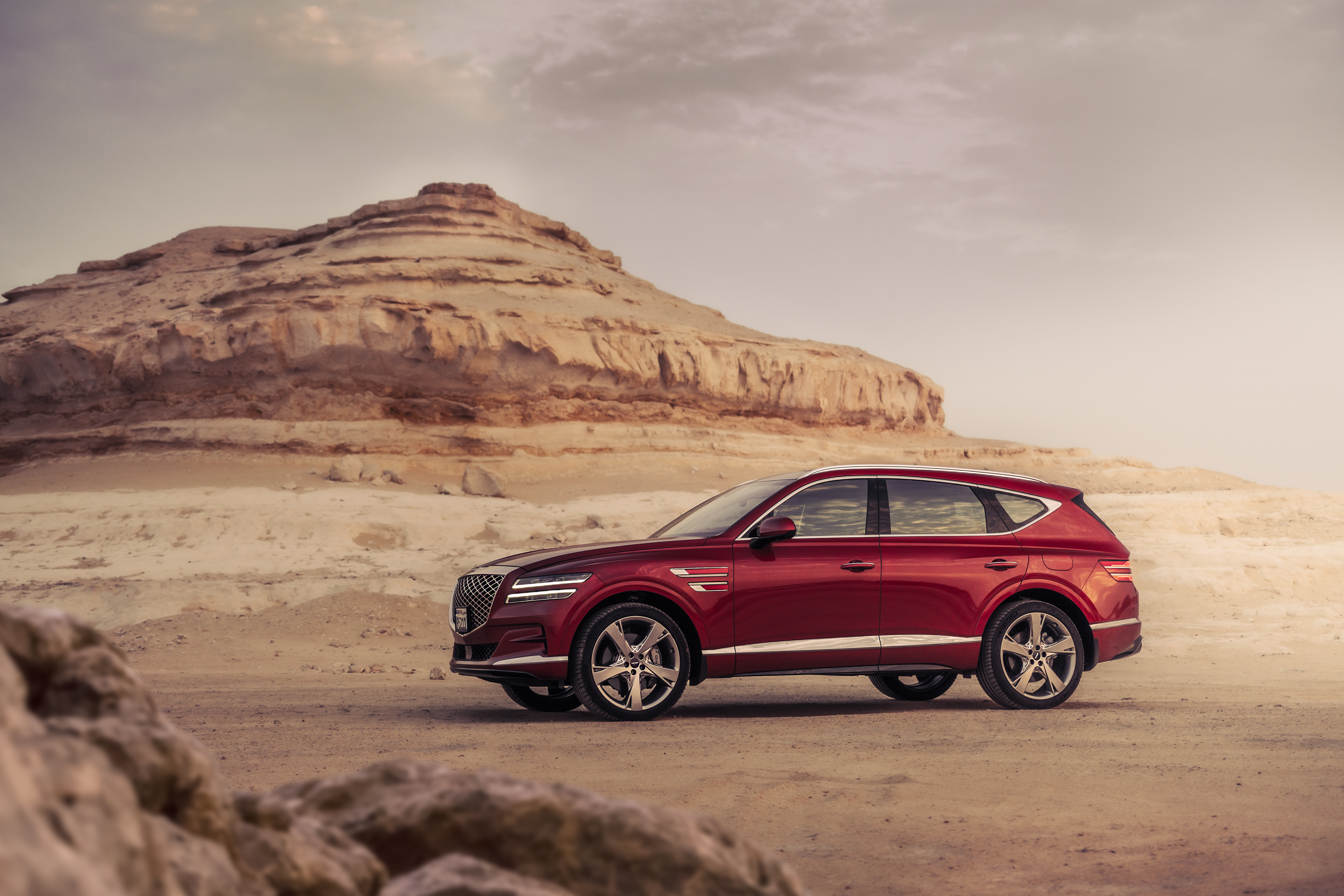 Side profile of Genesis GV80 driving through Bahrain’s Sakhir desert, soft morning light and sandstone ridges.