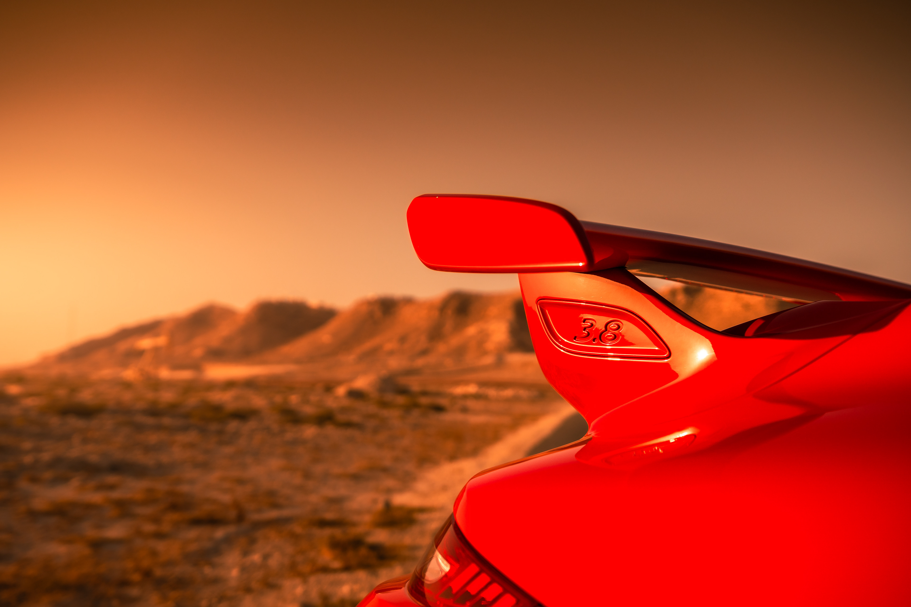 Close-up of the rear wing on a red Porsche 911 marked ‘3.8’ against desert hills at sunset.