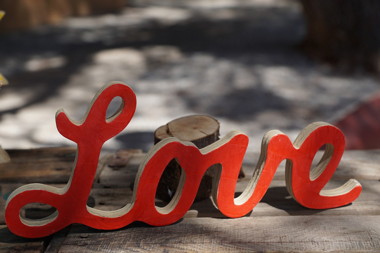 The word Love is a wooden cutout of cursive writing painted red sitting on a rustic wooden table and in front of a shaded path in the background