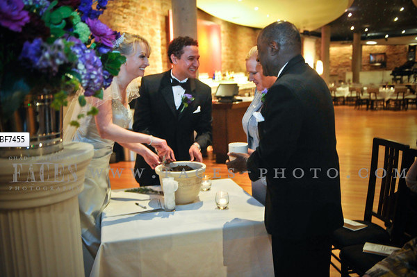 A bride in a cream wedding gown and a groom in a classic black tuxedo are standing around a table during their wedding ceremony with their parents and performing a Unity Seedling Ceremony by planting seedlings in a pot of soil