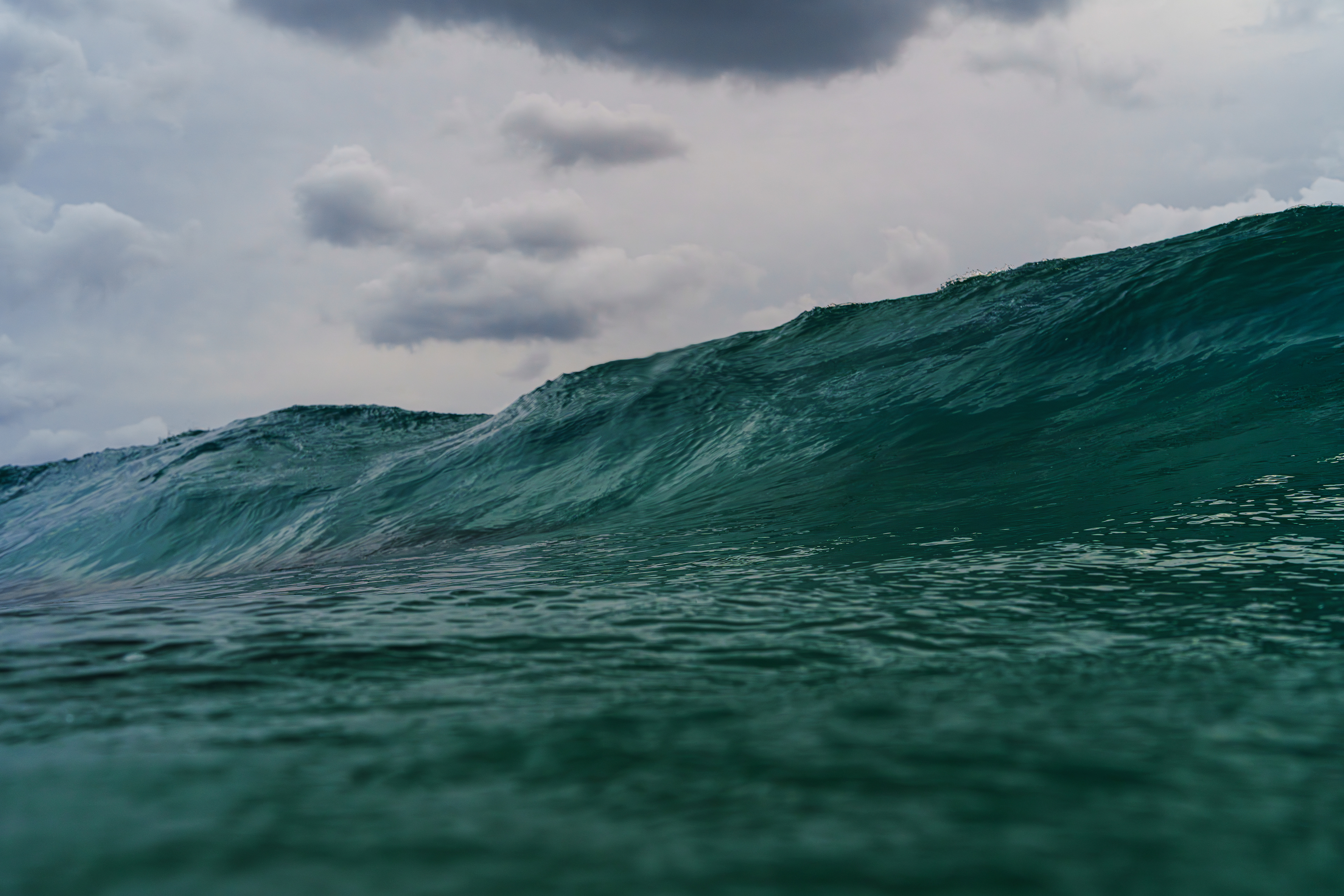 今年のTripは10日間、奄美大島に行く事になった。いつものメンバーとはタイミング合わずパートナーとキャンプ旅となった。もちろん撮影もするのでパタゴニアの100Lと60L,30Lのバックパックにキャンプ道具から撮影機材を詰め込み持って行った。キャンプ場は東シナ海にある有名なポイントの前。北風が吹くとサイズが上がる。たまたま行った日の2日前から冬型でキャンプ場に着いた夕方のときは胸肩サイズの波がブレイクしていた。明日の朝に期待をしてテントを設営して早めに眠った。翌朝、天気は悪く曇天でサイドオンの風が入り昨日ブレイクしていた波も消えていた。この風では太平洋側は期待できないと思ったが向かうことにした。到着すると少しジャンク気味だが肩〜頭のサイズ。6時が干潮で満ち込みは12時頃で見た感じでも浅いのが分かる。もちろん初めてのポイントなのでReefの状況も分からない。Reefに入るのは約20年ぶり...不安しかない状態。何台か波チェックをする車は来るが誰も入らない。時間が経つと風が強くなりそうだったので入ることに決めた。着替えてビーチに降りるとサイズがワンサイズ大きく見える。それ以上にReefの棚が黒くはっきり見える。これは浅い！エントリーしようとしていた場所を奥に移動した。遠浅である程度まで歩いて行くがビーチと違い強い流れで立っているのが難しい。ある程度の深さまで行って泳ぎ始めた。3つの棚があり初めの棚は潜れないほど浅い。何度も戻されながら何とかクリア。2つ目の棚は水深が少しあり何とか潜れた。それでも膝を何度か当ててしまった。アウトに出てセットが来ると黒い岩陰がはっきり見えてそこにヒットしてブレイクしていた。 2025/10/30　Amami Oshima  (1/1600sec F6.3 ISO800 40mm)