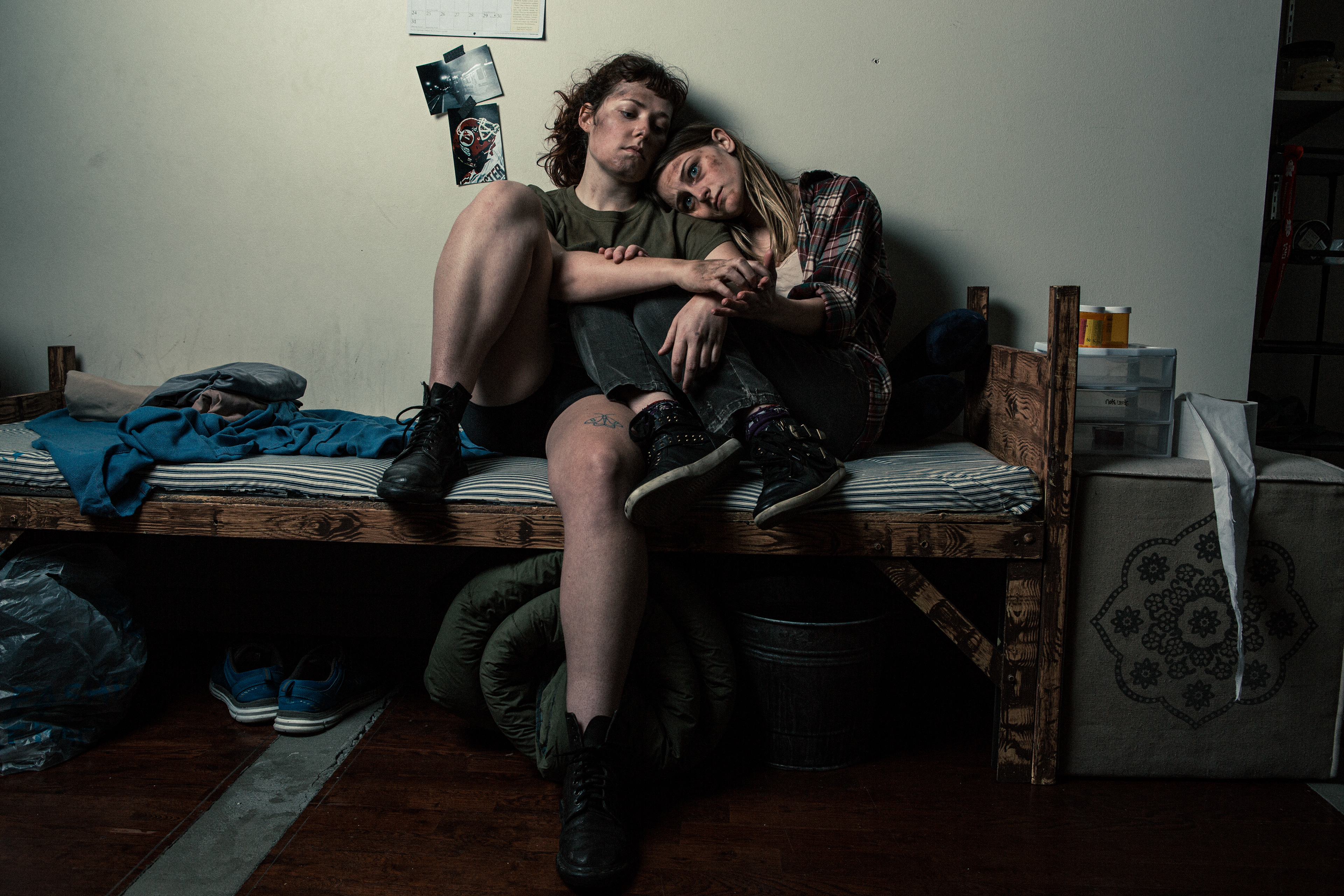 Resilient queer lesbian couple sharing a moment on a weathered bed within a modest dwelling. Their appearance reflects the challenges they've faced, with worn attire and tired expressions that hint at a difficult journey. The set, envisioned by Katherine Jelte, authentically captures the rawness of their experiences while highlighting the strength of their connection.