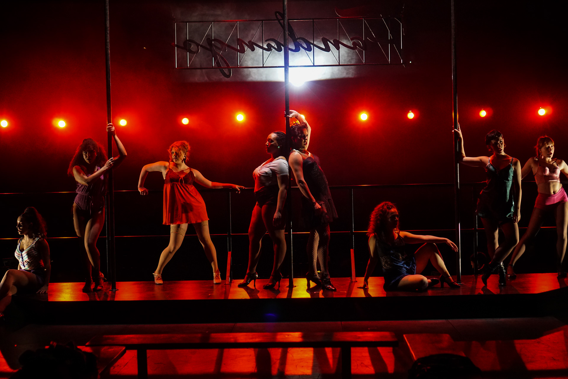 Vibrant scene on a theatrical stage featuring dancers in elegant poses beside gracefully adorned poles. The dancers' forms are softly illuminated by the warm glow of crimson lights, casting a passionate ambiance. The backdrop showcases a sign reading 'Fandango,' adding to the allure of the performance. This captivating set design, brought to life by Katherine Jelte, exudes an air of sophistication and artistry.