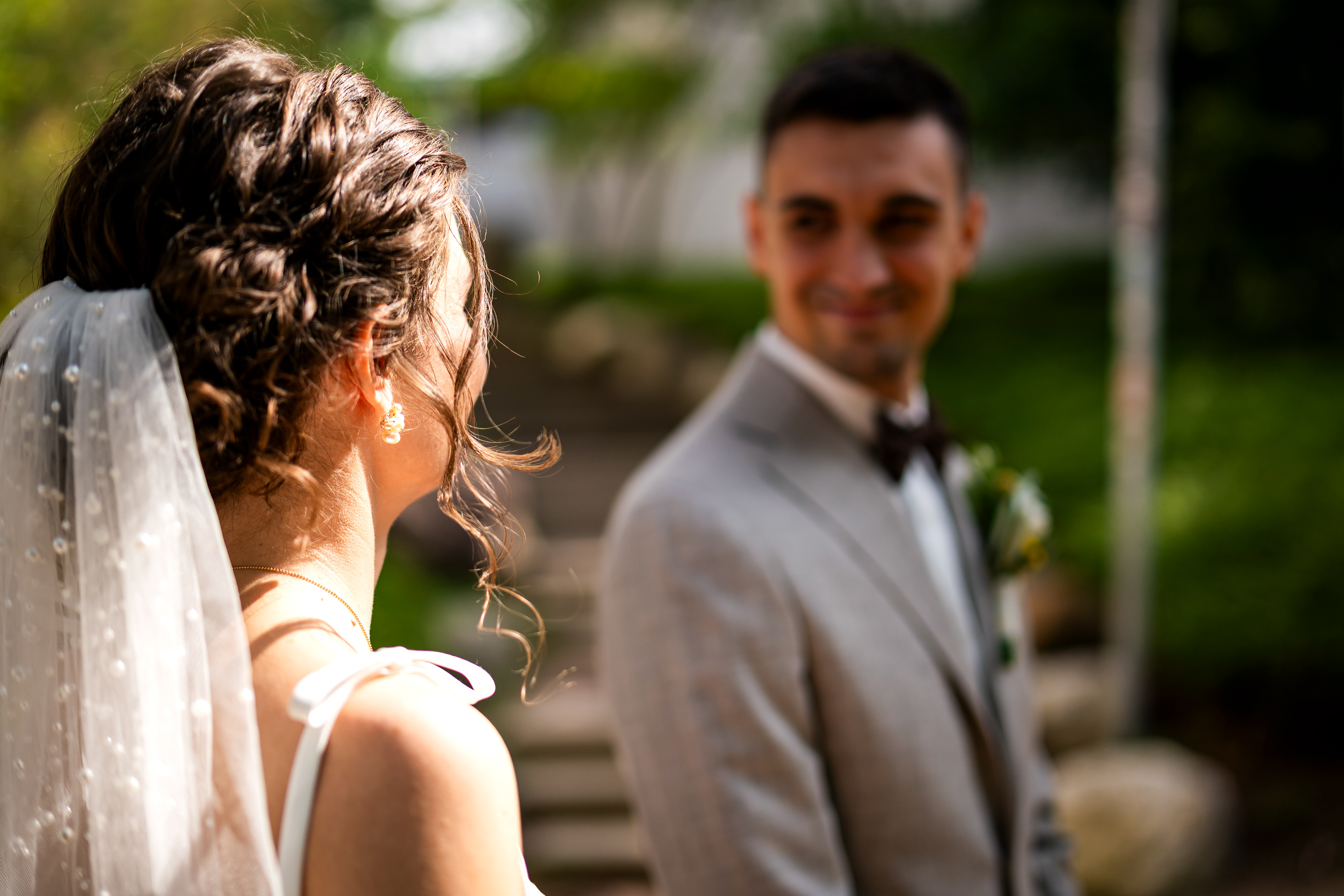 Braut Melanie im Vordergrund blickt lächelnd zu ihrem Bräutigam Simon im Hintergrund. Natürliches Hochzeitsporträt aus der Reportage von MFUCHSLOCH Fotografie & Videografie.