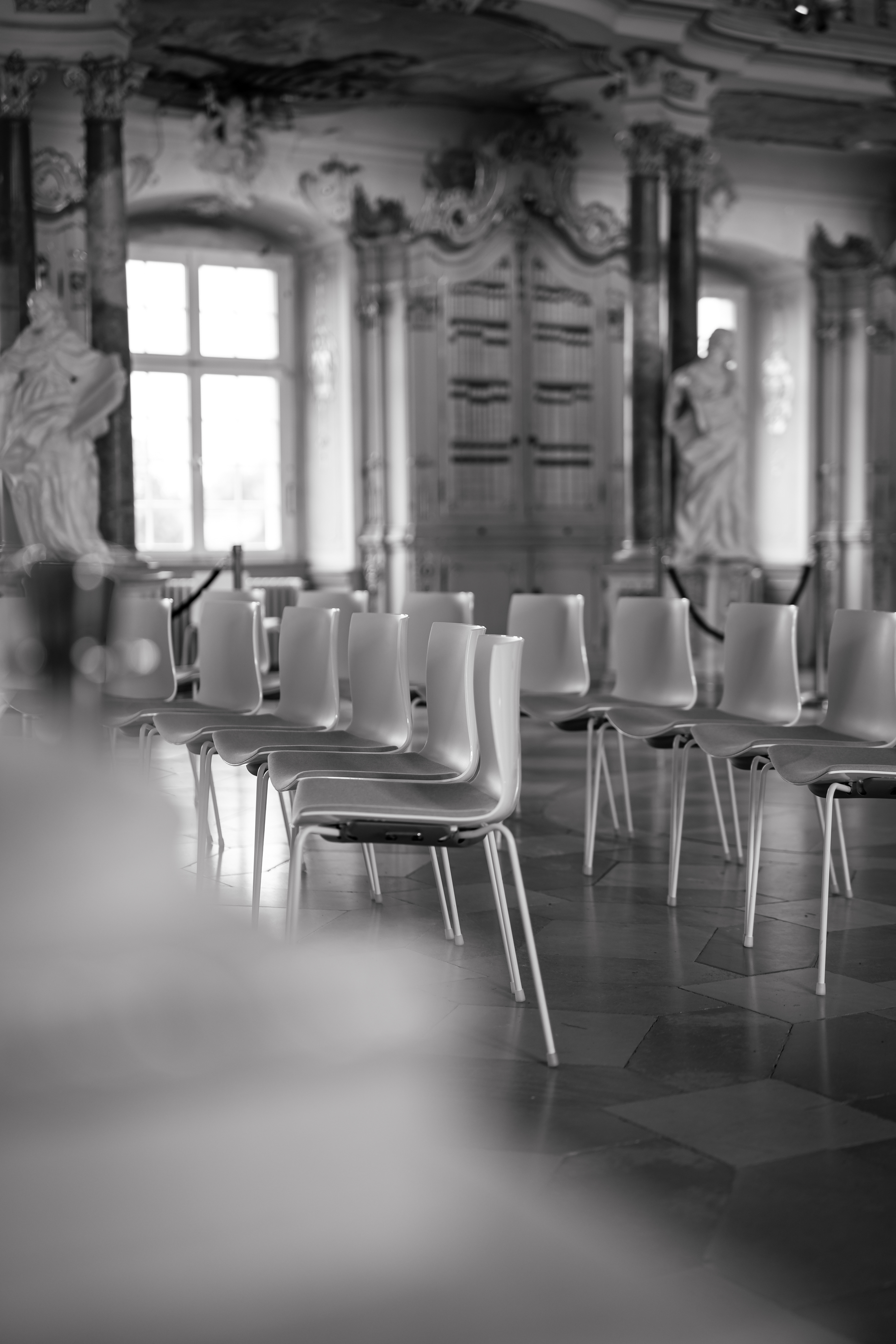 Atmosphärische Schwarz-Weiß-Aufnahme der leeren Gästestühle kurz vor Beginn der standesamtlichen Trauung. Künstlerischer Fokus auf die Details im historischen Bibliothekssaal Bad Schussenried. Dokumentarische Hochzeitsfotografie in Oberschwaben von MFuchsloch.
