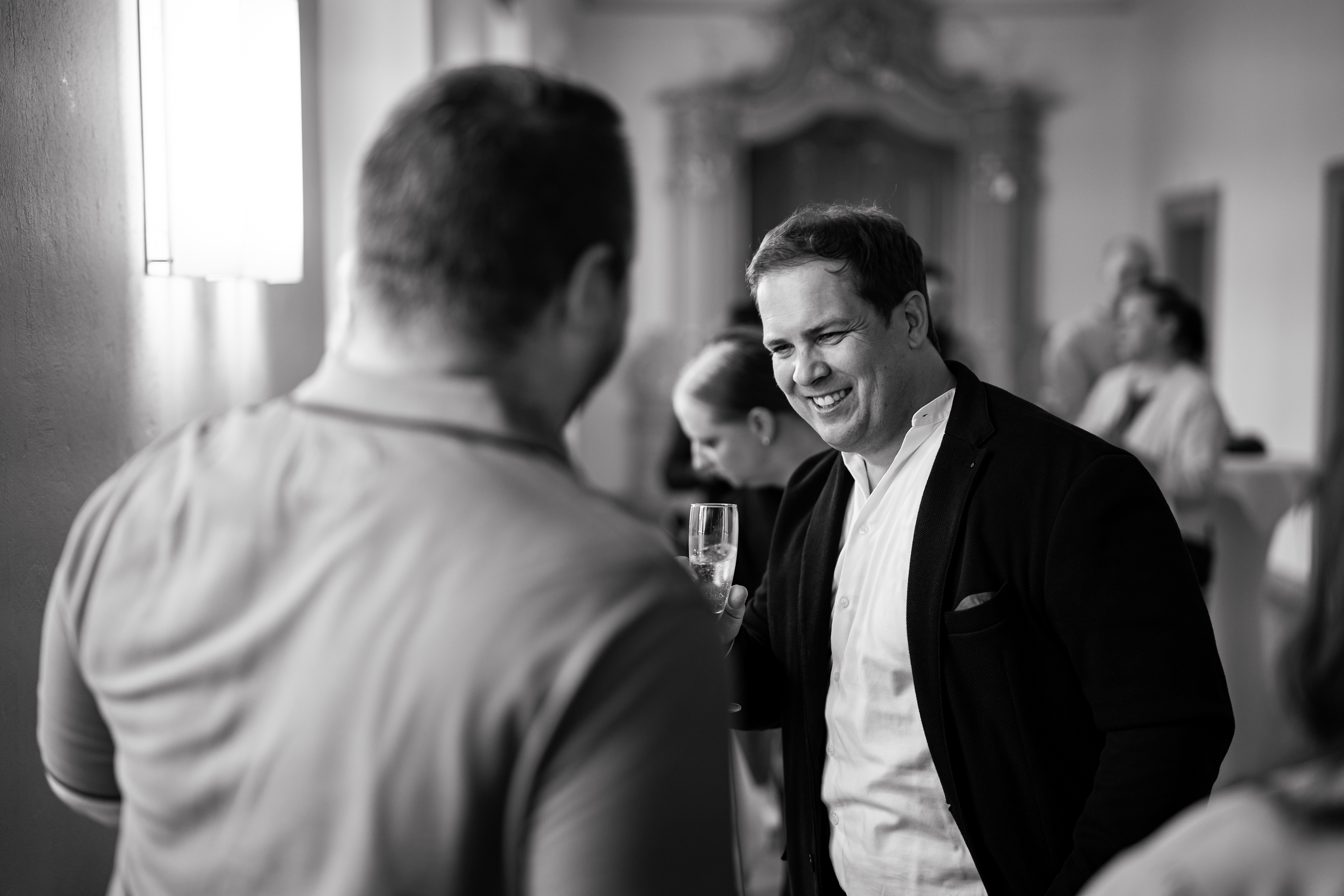 Bräutigam Dominik lacht entspannt beim Sektempfang mit Gästen im Kloster Schussenried. Schwarz-Weiß Hochzeitsreportage von MFUCHSLOCH.