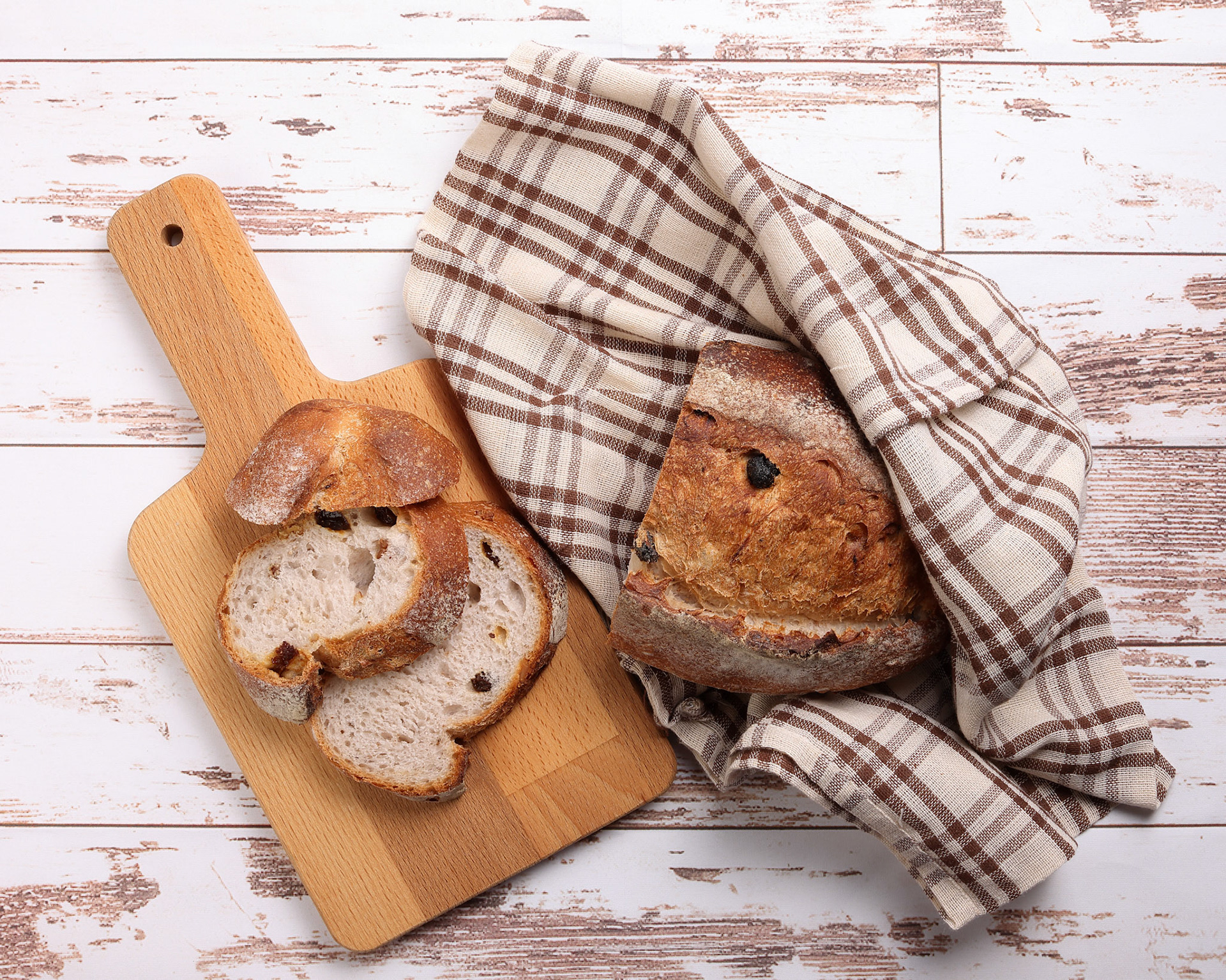 Rustic whole grain artisan bread loaf slice with cranberry raisin dry fruit nuts wrapped in checkers cloth with wooden chopping board over table top flat lay view