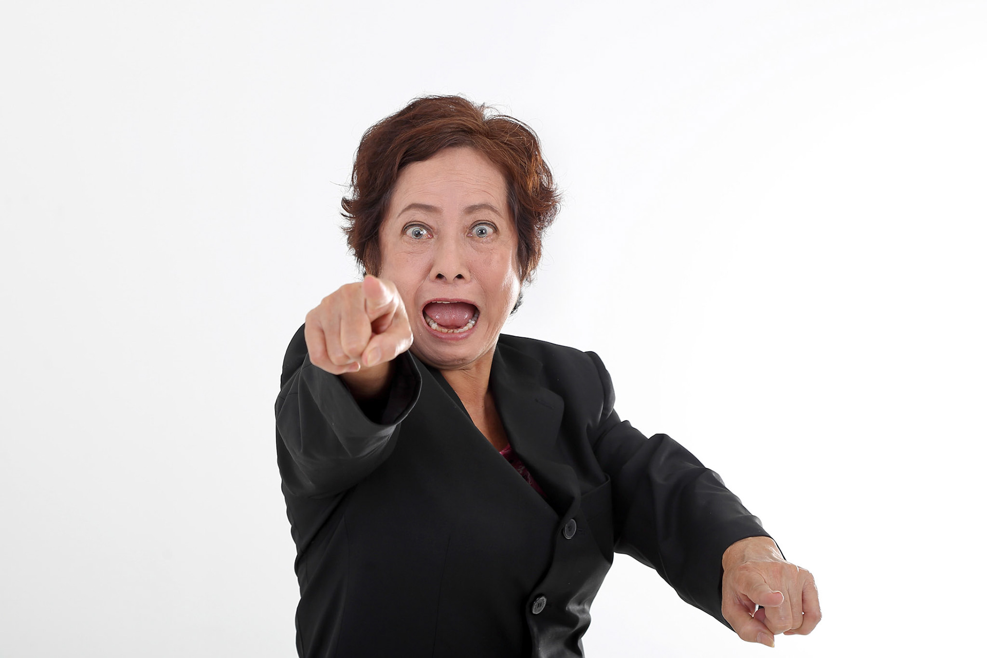 Elderly asian Chinese female on white background wearing black suit point at camera angry furious mouth open shout