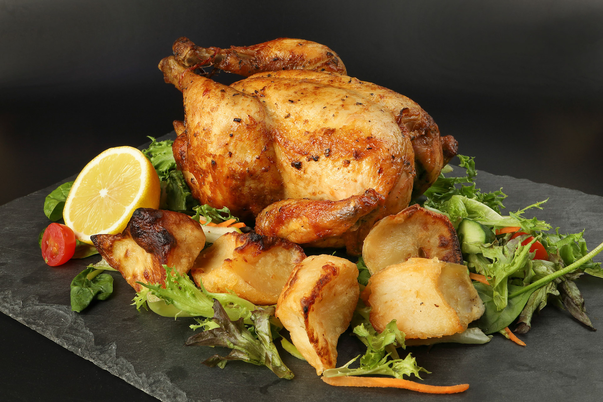 Whole roasted grilled chicken poultry bird with baked potato vegetable salad tomato lemon on black slate stone cutting board black background