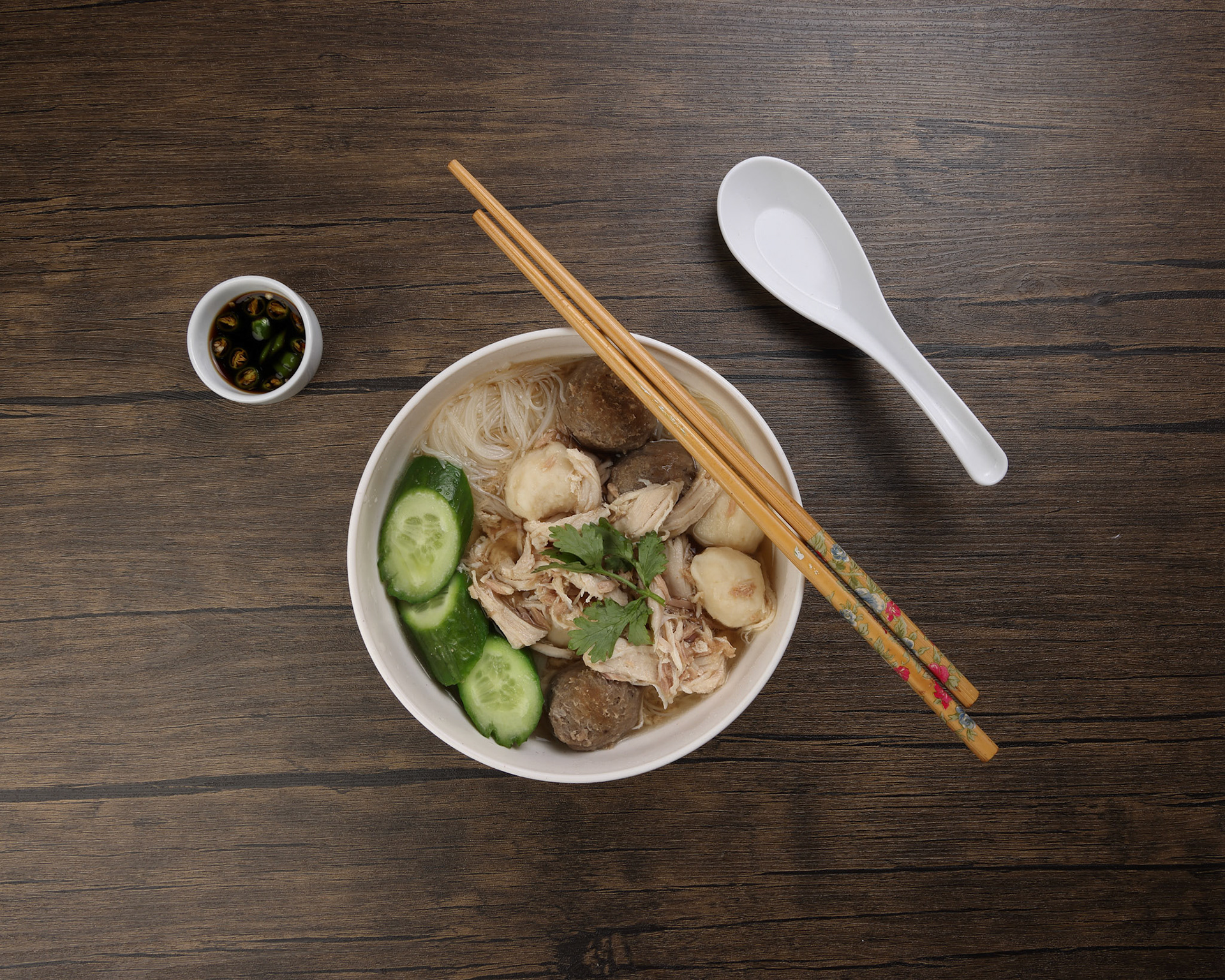 Clear white rice vermicelli beehoon noodle soup with chicken beef meat ball shredded cucumber in white bowl chopsticks spoon chili soya sauce parsley garnish