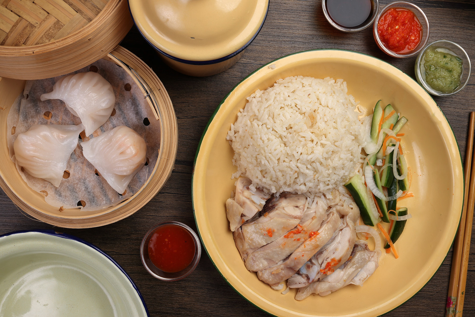 steamed Hainanese chicken rice crystal har gow transparent prawn shrimp meat stuffed dim sum in bamboo steamer green red chilli ginger soya sauce cucumber carrot on retro enamel diner plate fork spoon chopsticks on wooden table