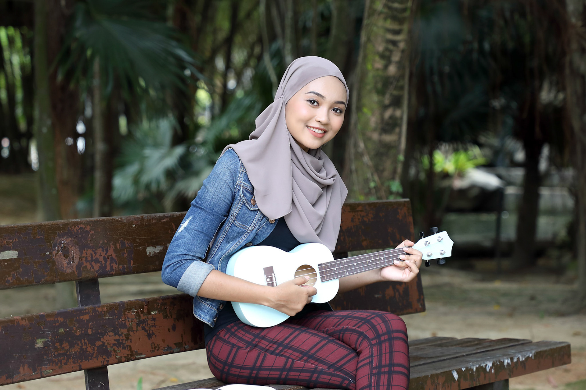 Young Asian Malay woman headscarf outdoor green sit on park bench play ukulele music sing