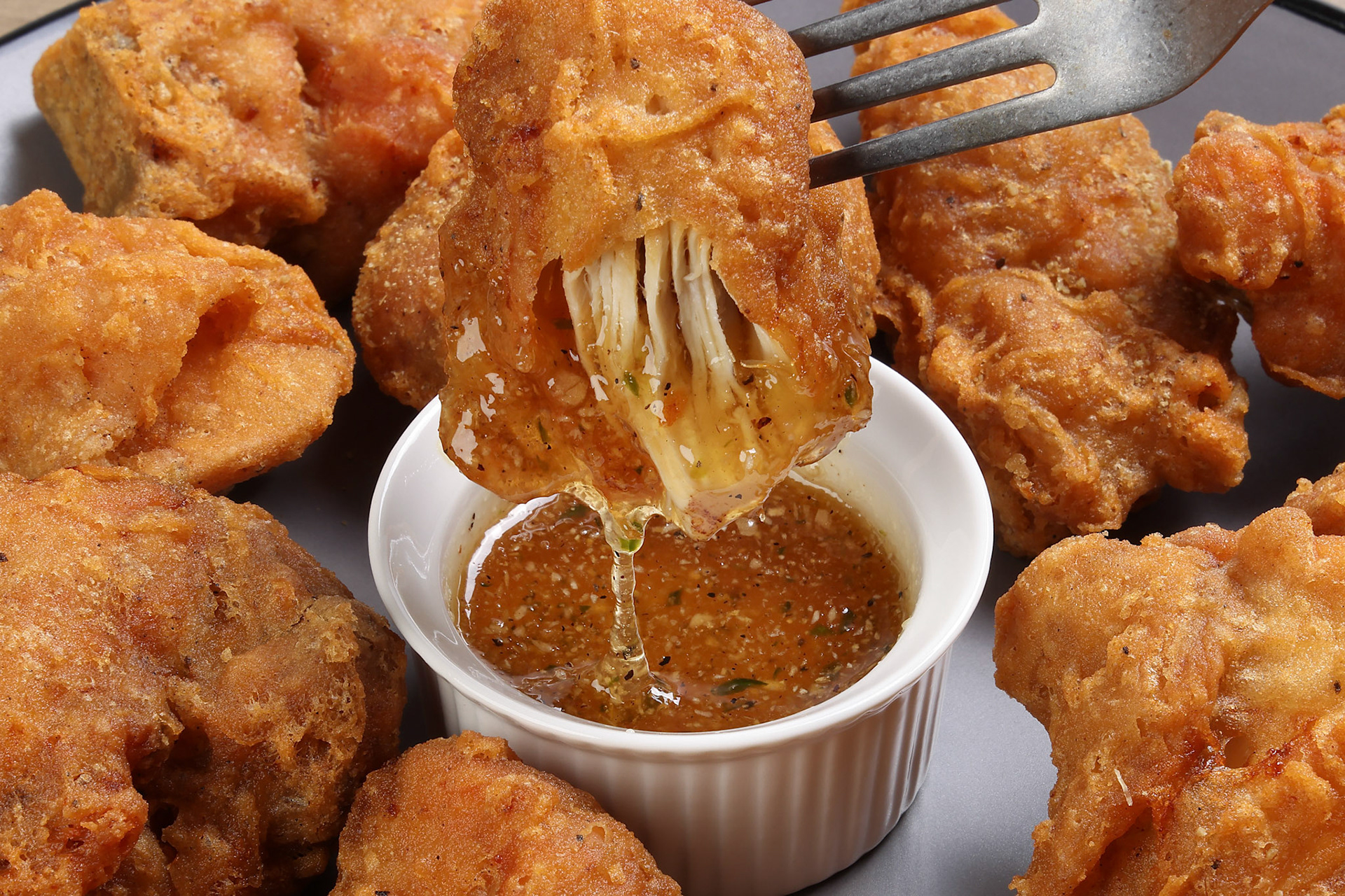 Light battered deep fried chicken variety mix pieces pile flaky meat on grey plate on fork dripping honey garlic clear sauce in center over wooden background