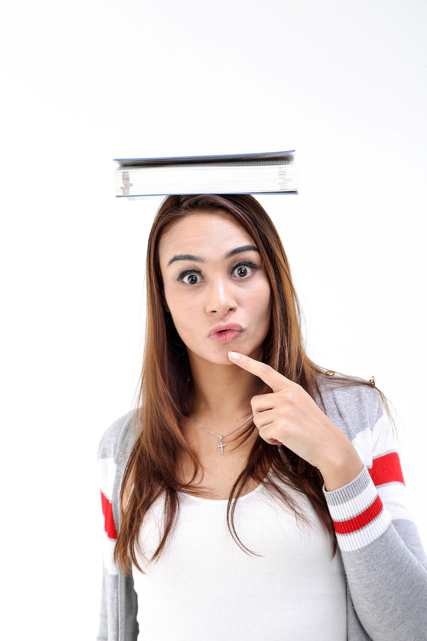 Yong Asian youth girl in casual dress balancing book on head look at camera finger on chin expression think shocked funny on white background