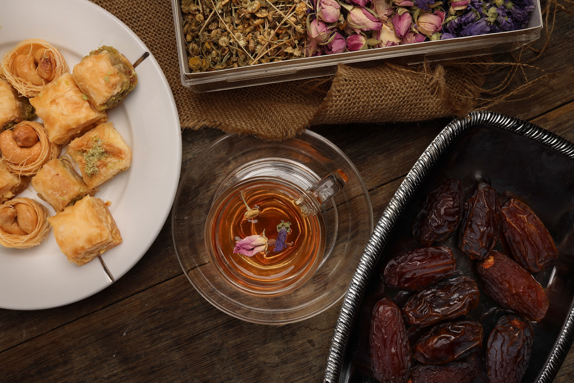 Layered stuffed pastry Baklava middle eastern sweet dessert dry ripe sweet date fruit dry flower tea rose bud butterfly pea chamomile flower tea in transparent glass cup saucer on rustic wooden background