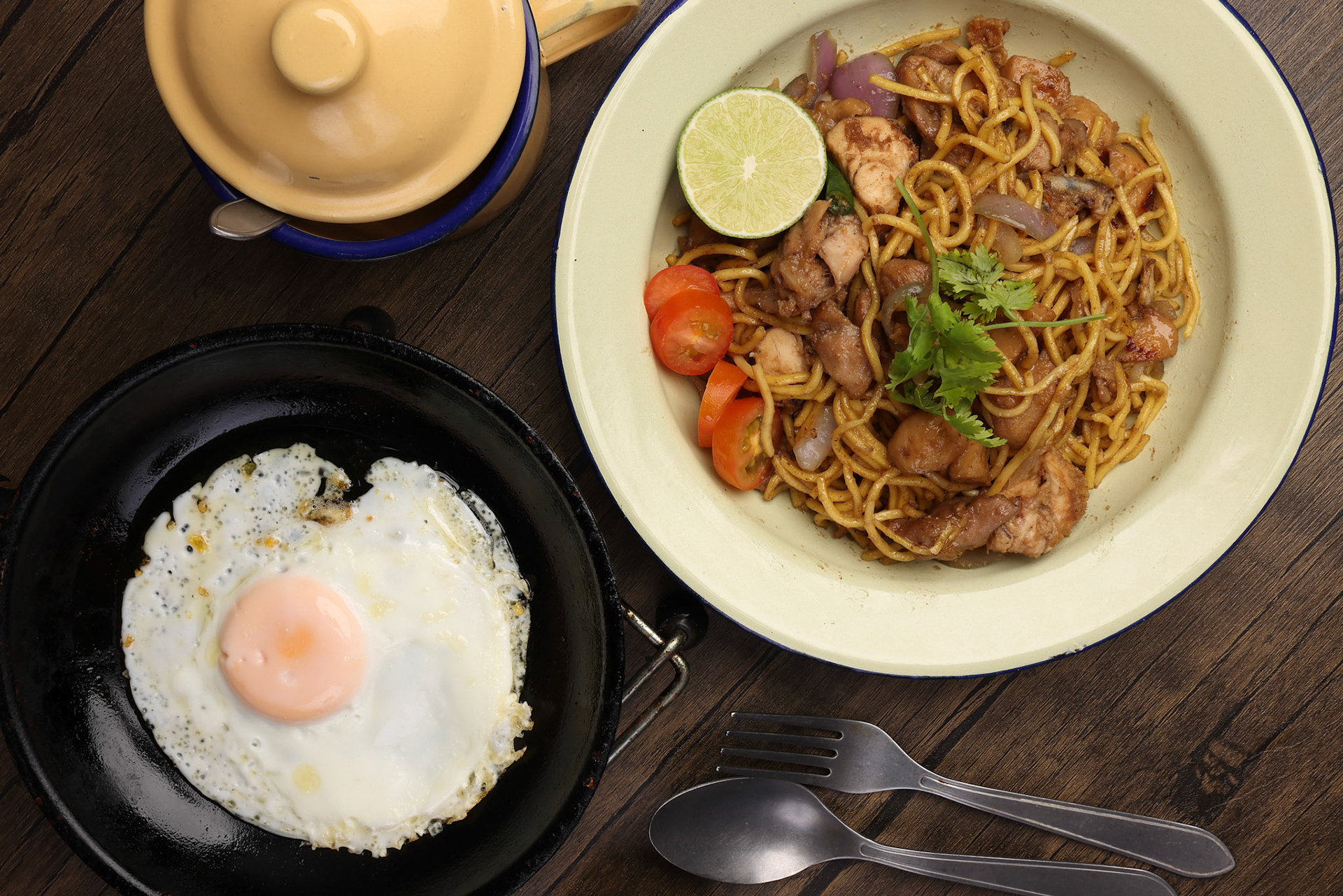 Stair fried yellow noodle traditional mee gorng tomato parsley lime on enamel metal plate cup sunny side up egg in frying pan fork spoon dark soya sauce
