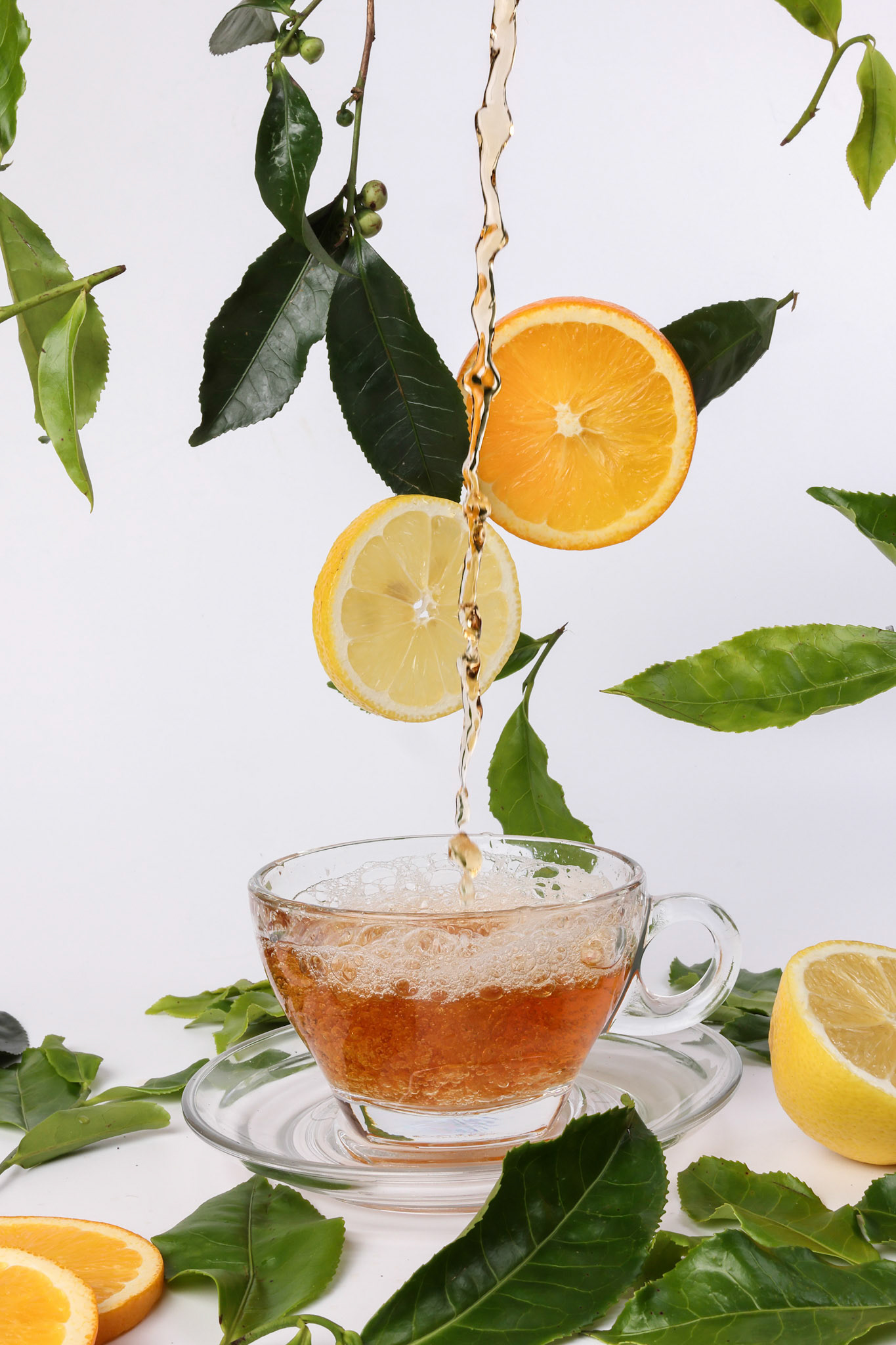 different types of fresh raw green tea leaf flower bud dropping floating elevated lemon orange slice over transparent glass teacup saucer pouring dripping liquid tea on white background