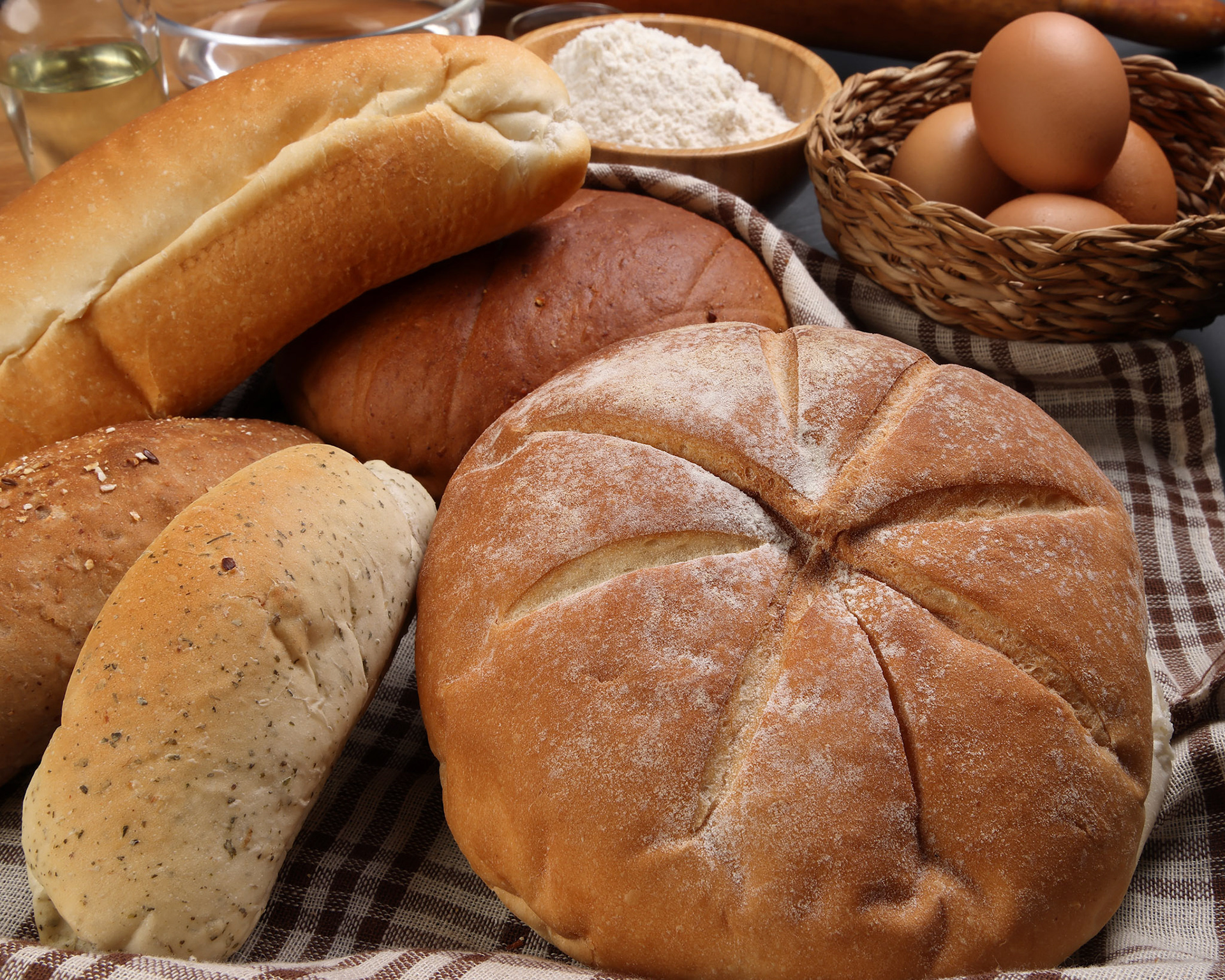 Freshly baked bread loaf bun roll round long mix verity wrapped in checkered kitchen fabric napkin towel wheat flower oil water salt eggs rolling pin slate stone over black background