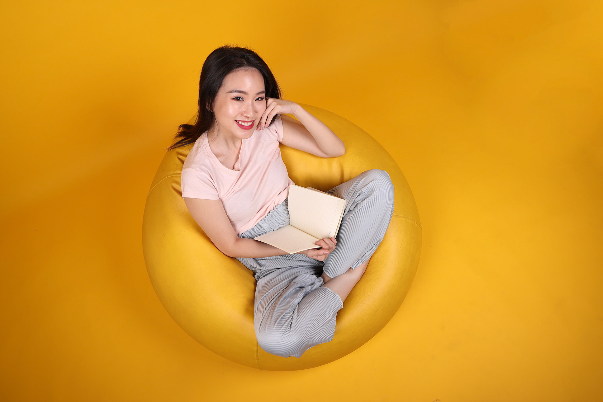 Beautiful young south east Asian woman sits on a yellow beanbag seat orange yellow color background pose fashion style elegant beauty mood expression rest relax think emotion read book top view