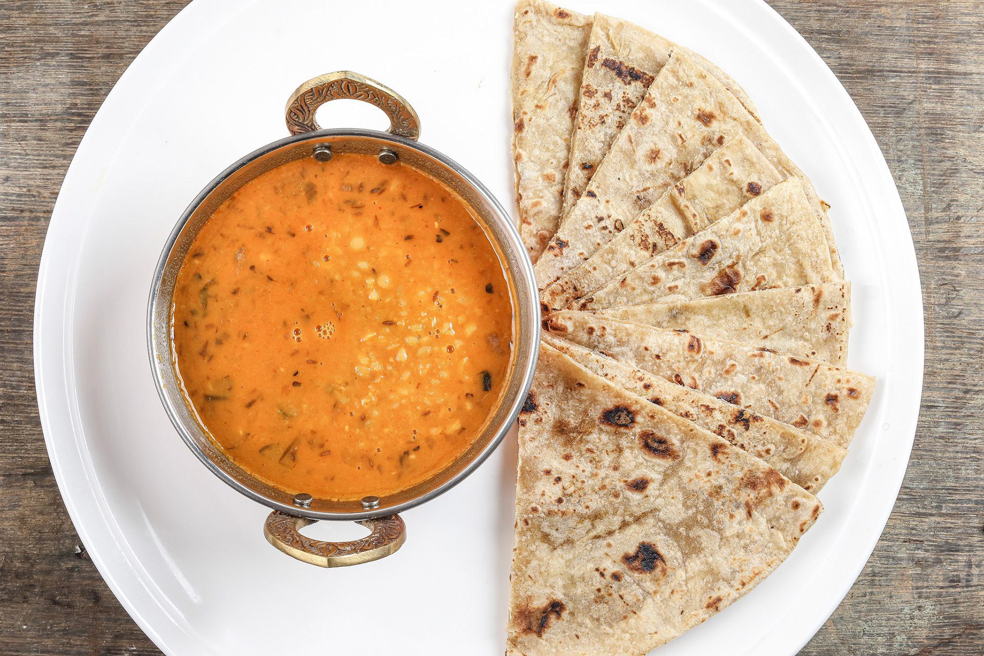 Handmade flat bread chapati and dal tadka fry in brass bowl pan on white plate rustic wooden background